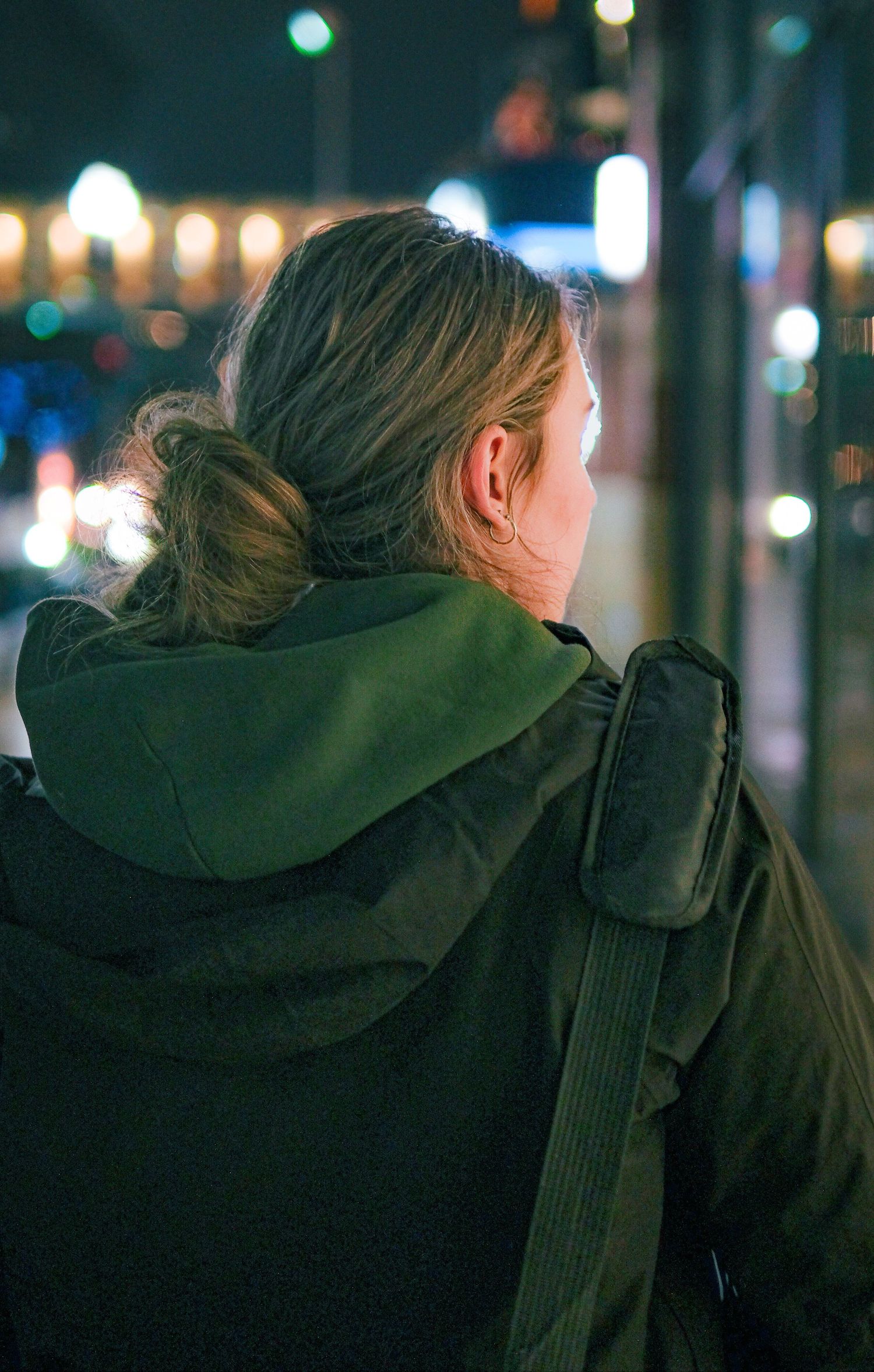 Person in a dark green hooded jacket looking away at night with city lights blurred in the background.