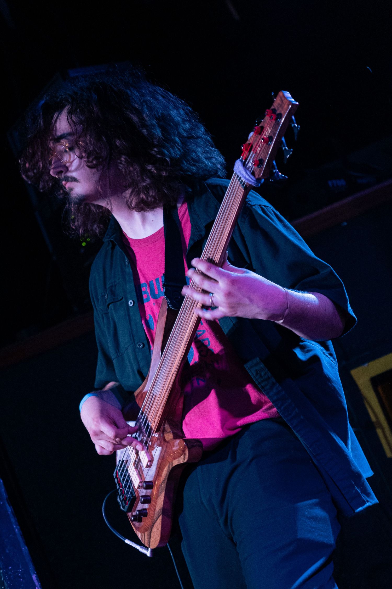 A musician in a dark jacket and pink shirt plays bass guitar on stage under dramatic blue lighting.