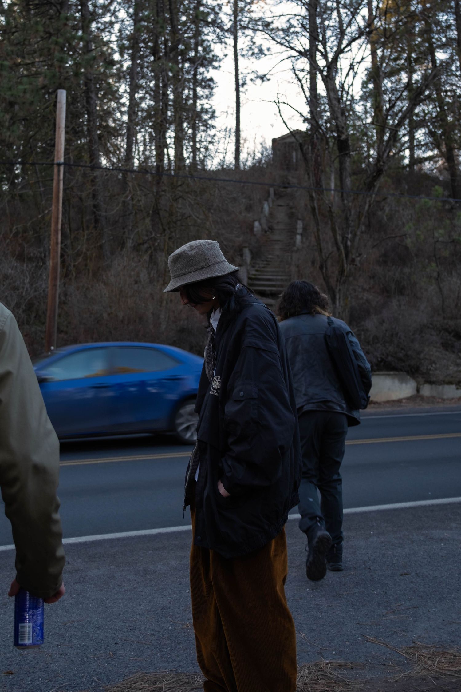 Several people in dark winter clothing stand near a road at dusk with a blue car visible in the background.