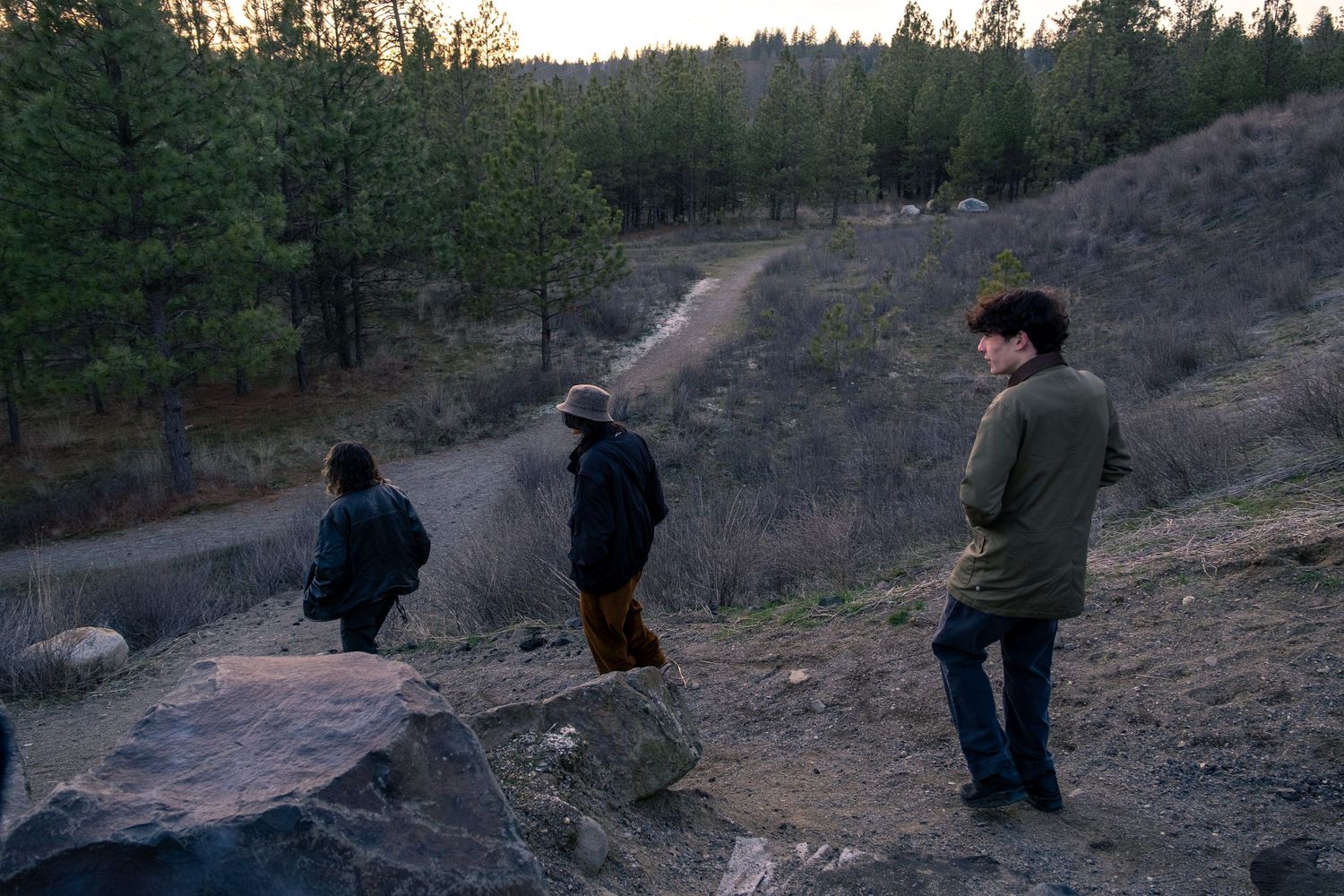 Three people standing on hillside overlooking trail in wooded area at dusk.