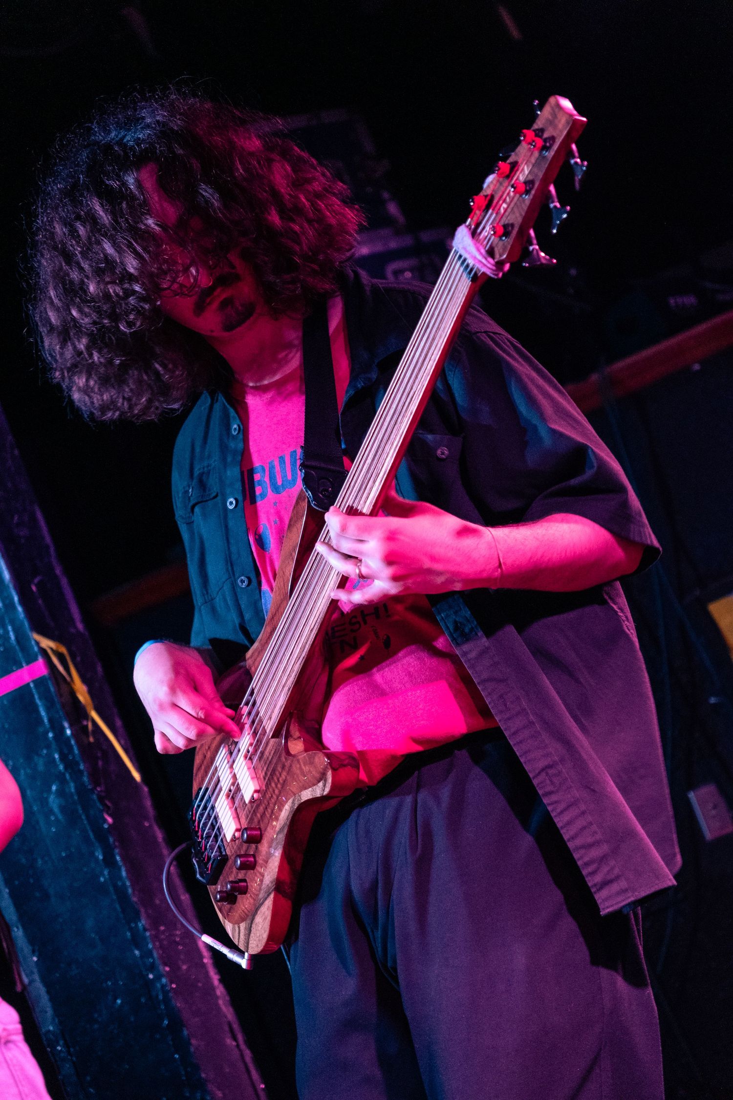 Musician in dark clothing plays bass guitar under vibrant pink stage lighting during a live performance.