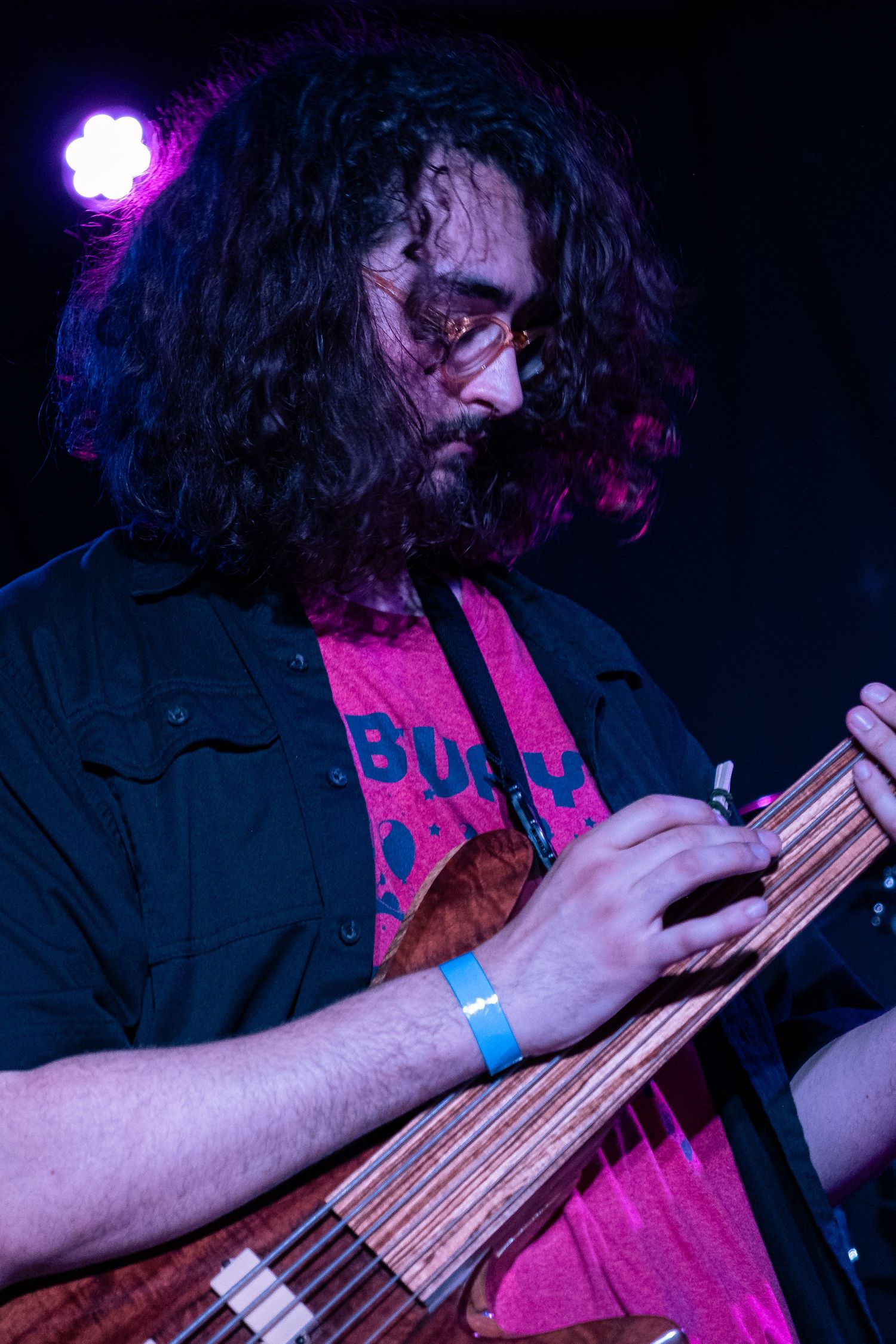 A musician plays fretless bass guitar in dramatic purple and blue stage lighting during a performance.