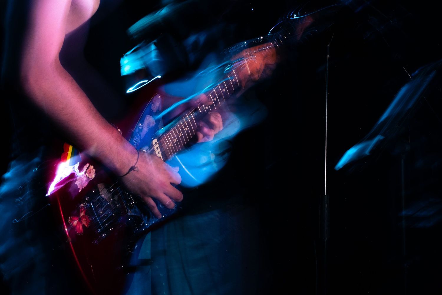 A guitarist plays an electric bass guitar on stage illuminated by dramatic blue and pink lighting during a performance.