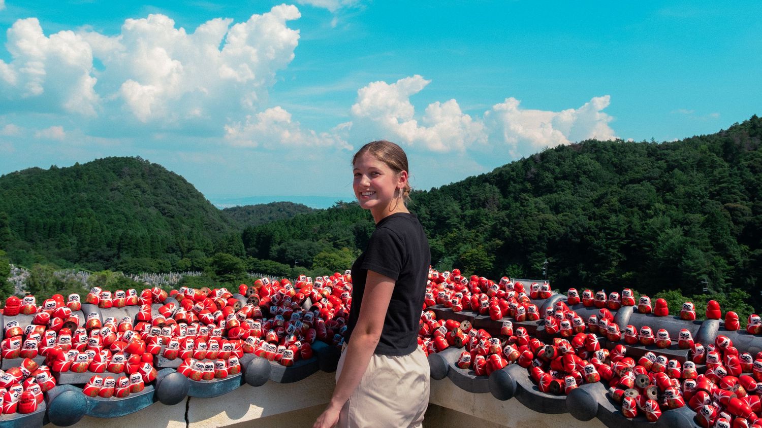 A panoramic view of red kayaks lined up along a shoreline with green hills and blue sky in the background.