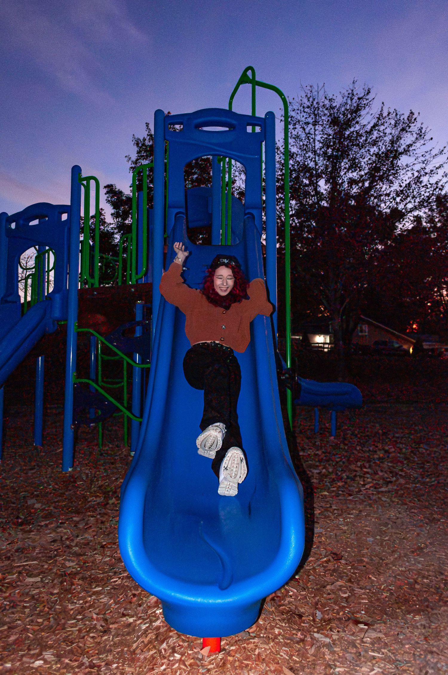 A sequence of five photos showing someone sliding down a blue playground slide at dusk with autumn leaves on the ground.