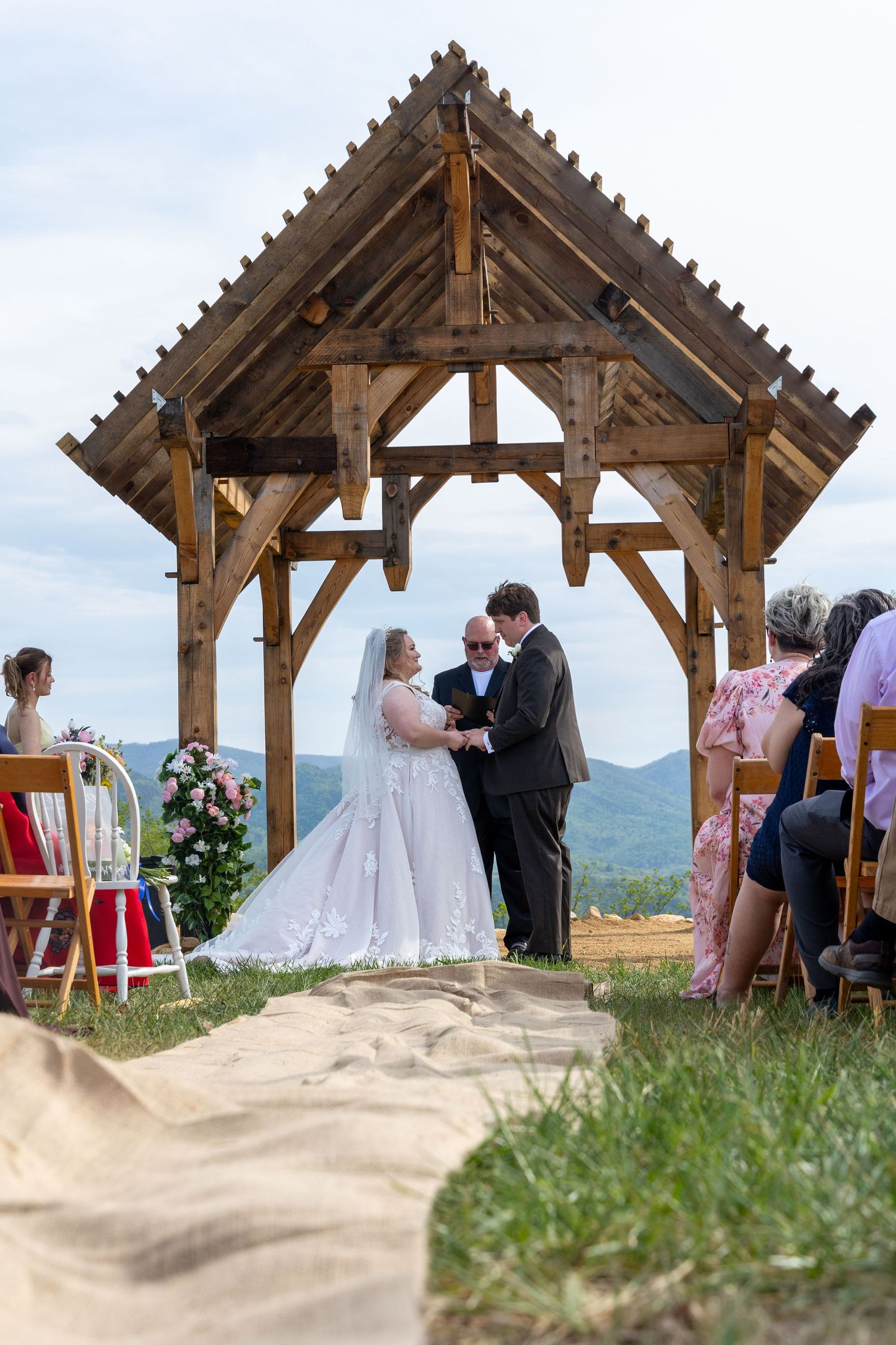 Bride and groom exchange vows under a rustic wooden pavilion at an outdoor mountain wedding ceremony with guests seated nearby.