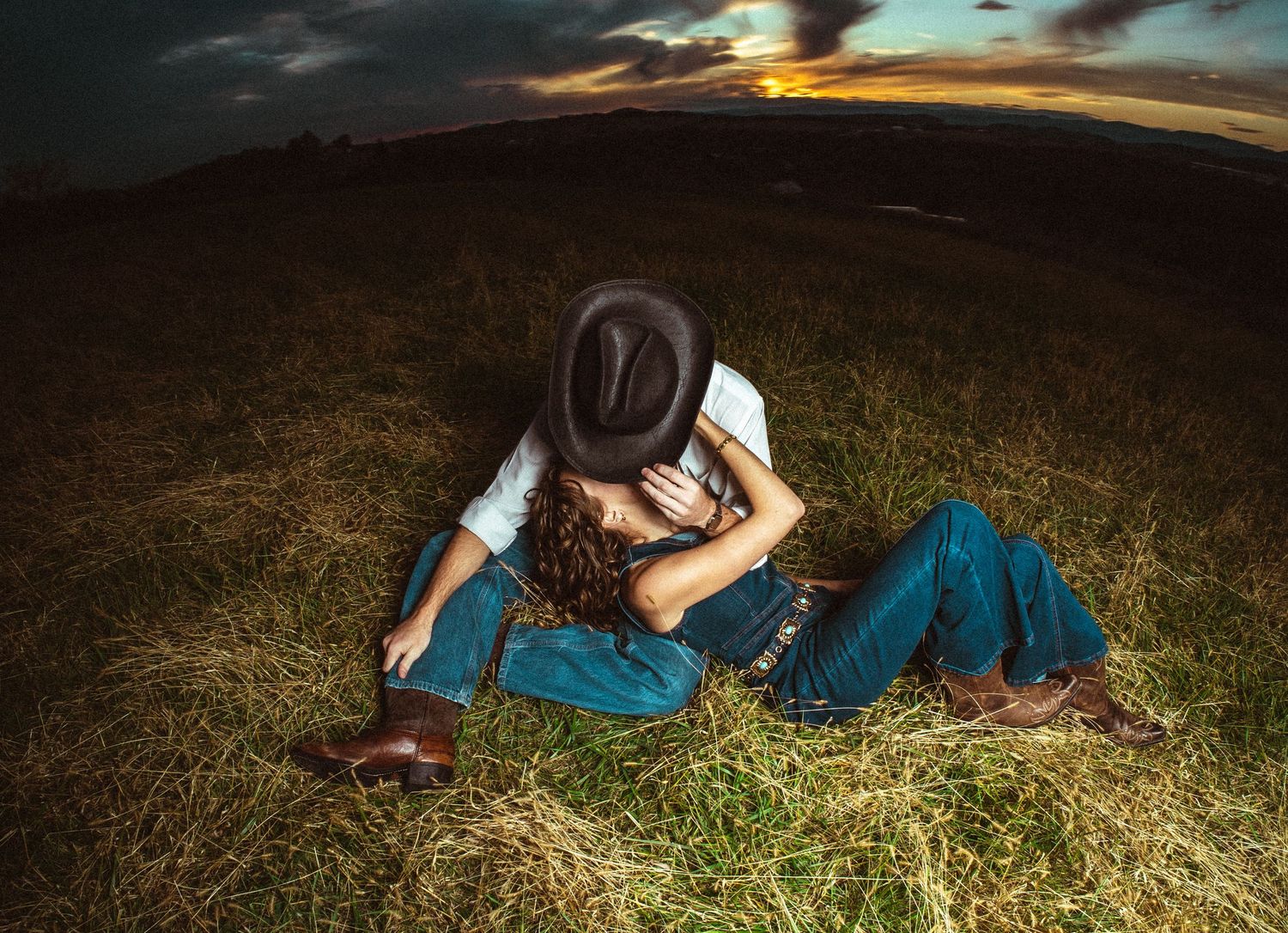 Two people in denim and western wear embrace while lying in a grassy field at sunset.