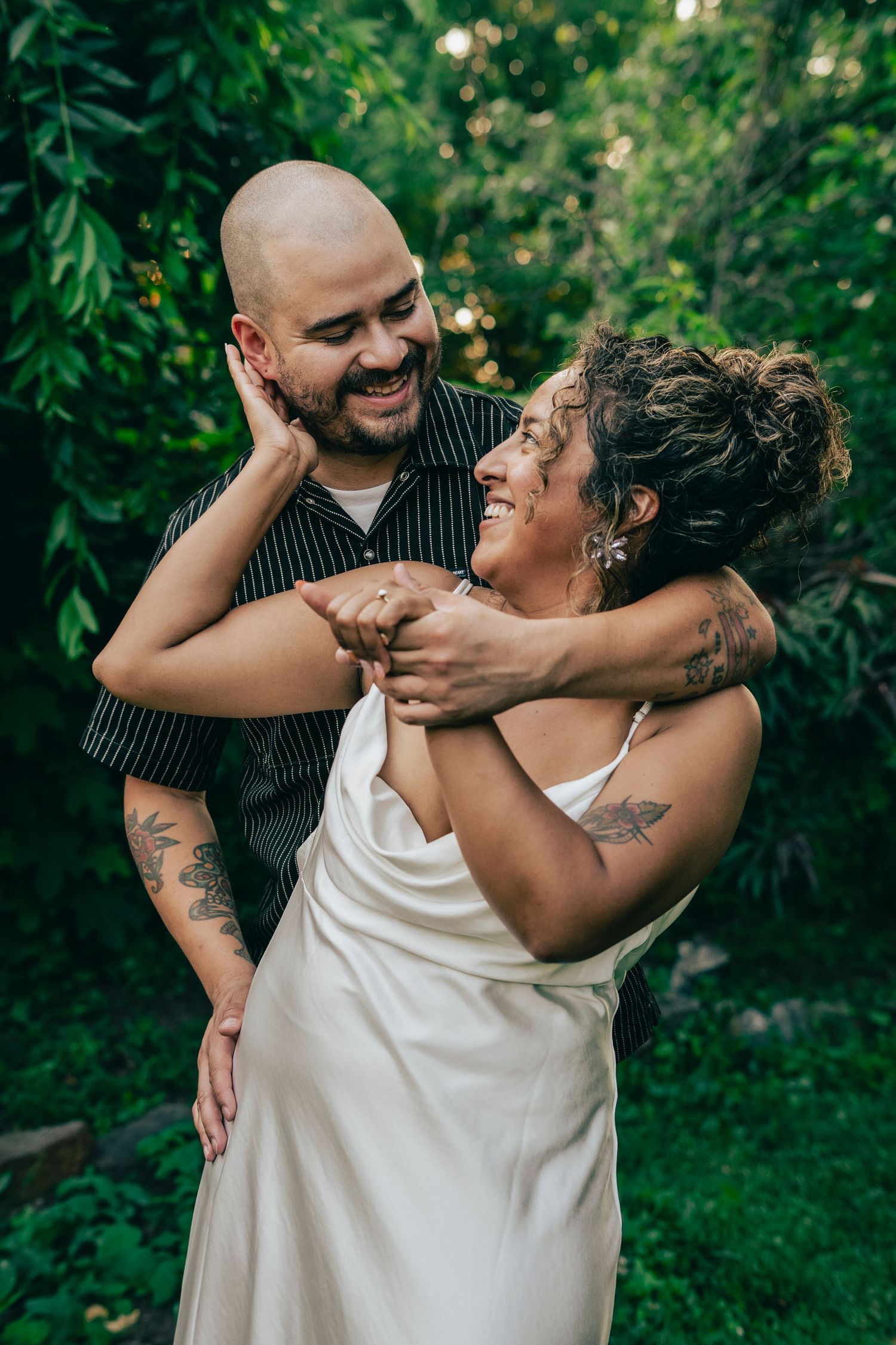 A couple shares an intimate moment dancing and laughing together in a lush green garden setting.