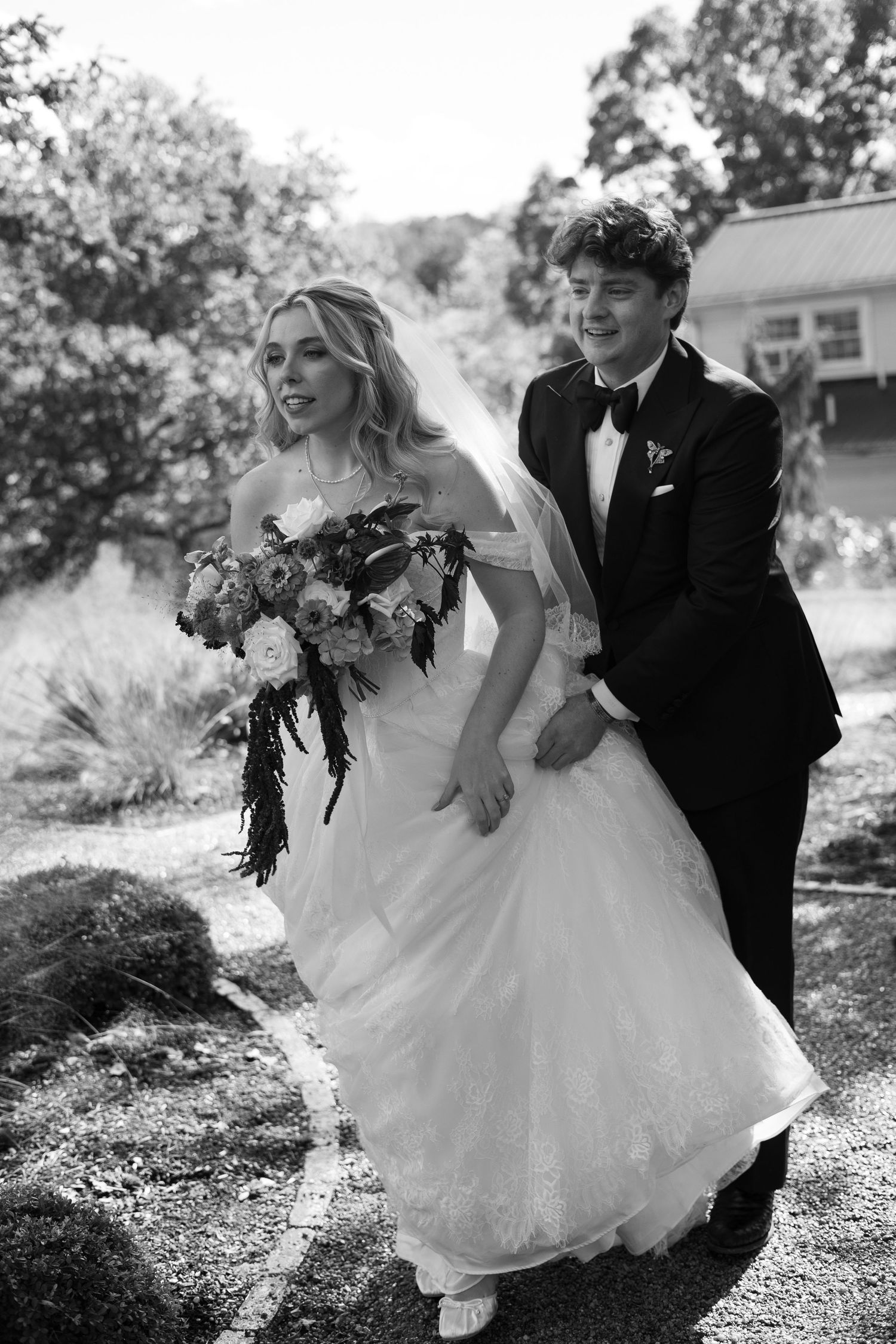 Black and white photo of a bride holding a bouquet and a groom in a tuxedo and bow tie walking together outdoors on their wedding day.