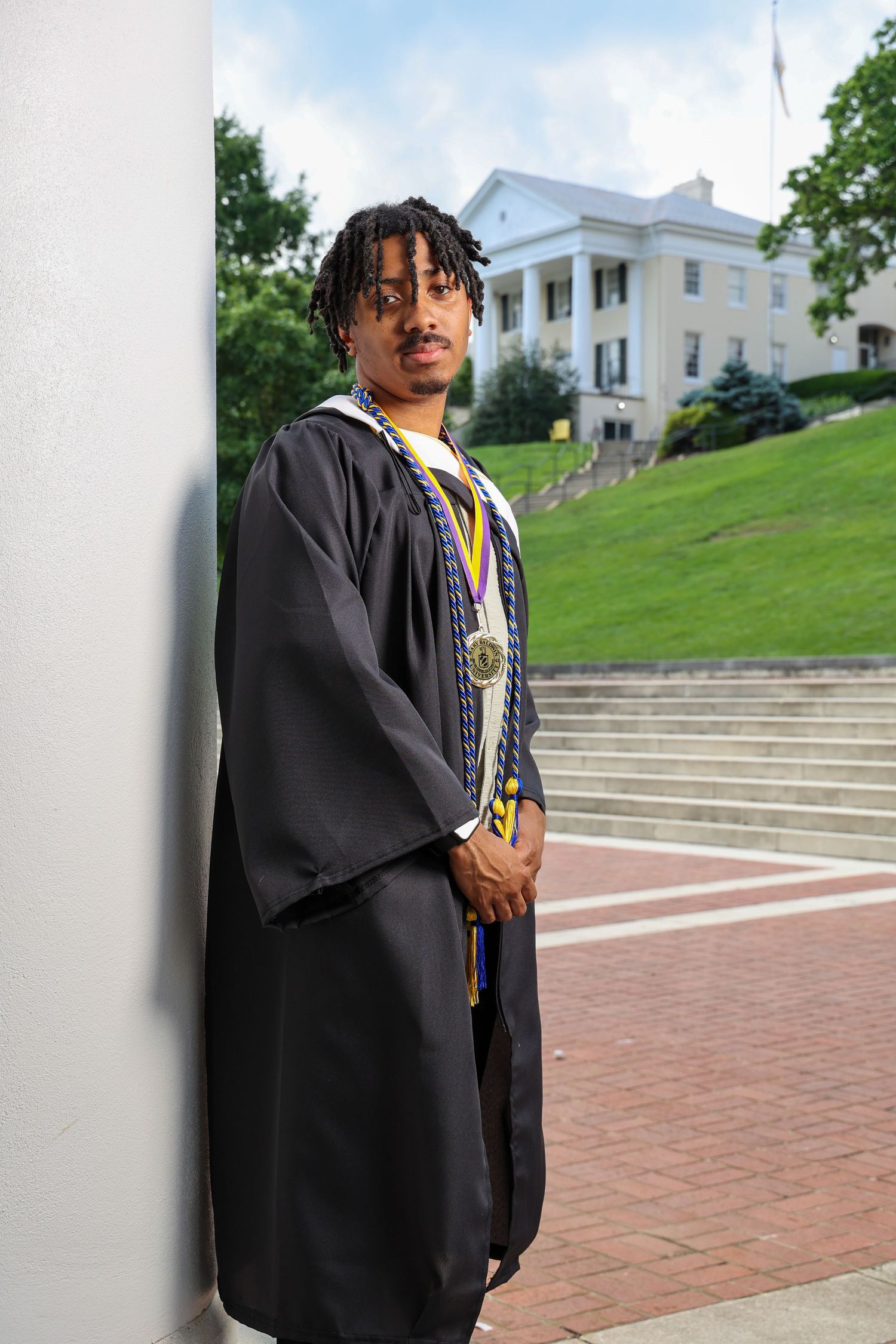 Graduate wearing black academic regalia poses against white columns on a brick courtyard with stairs in background.
