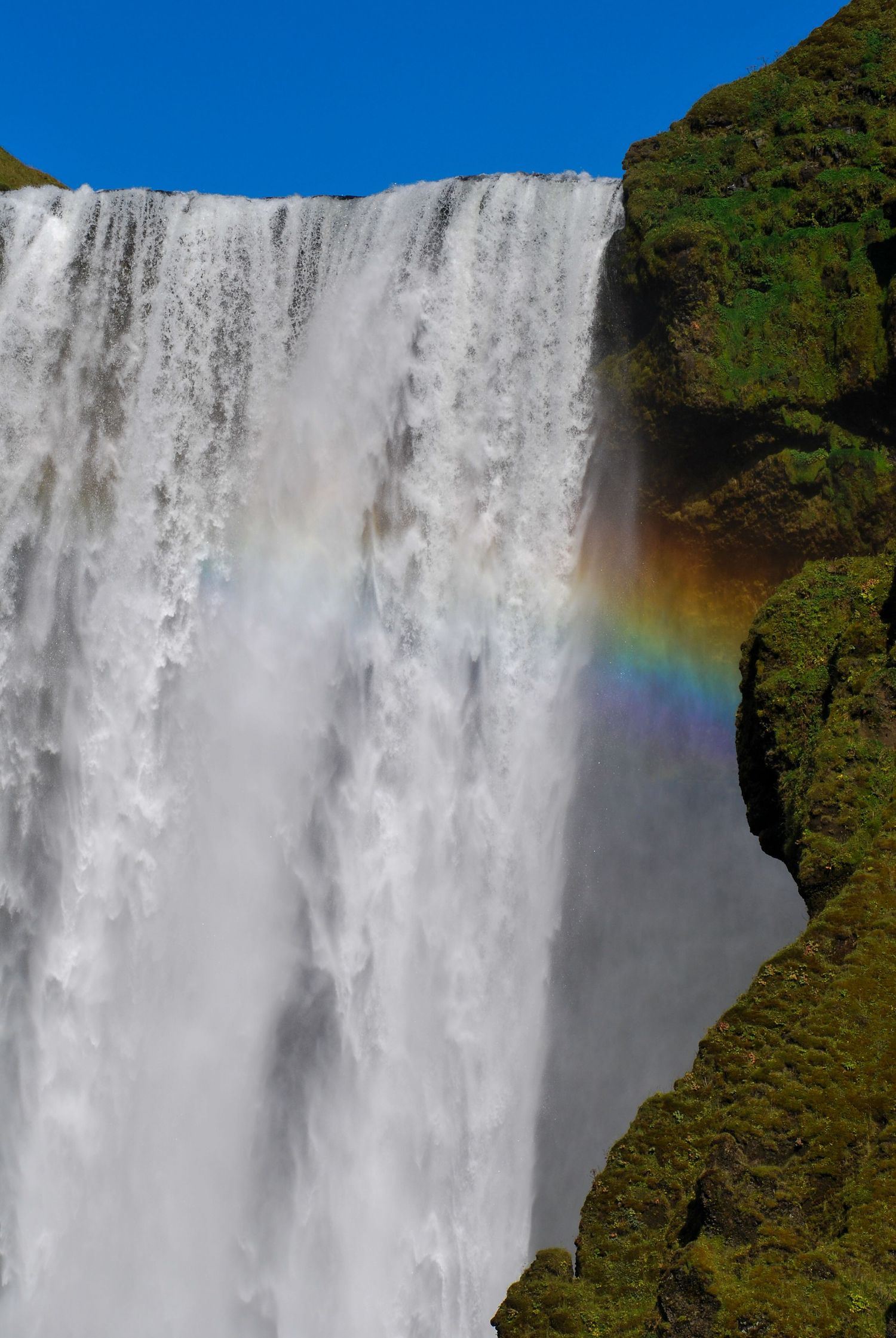 Majestic waterfall with rainbow mist against blue sky and green vegetation.
