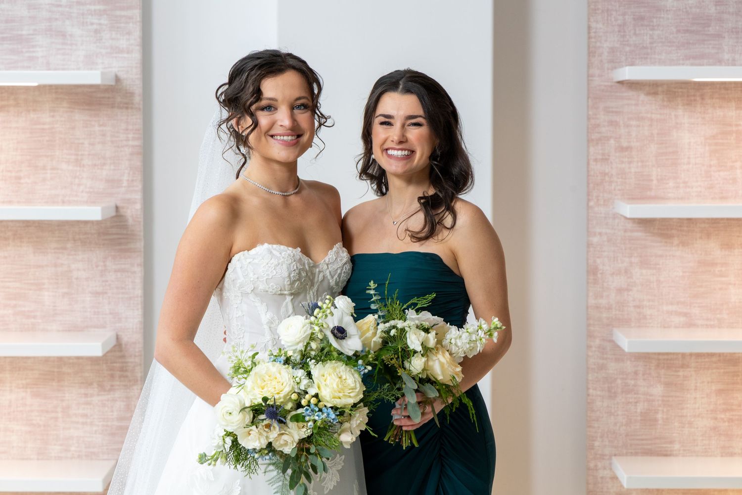 Two people in wedding attire pose together with white floral bouquets against a light colored wall.