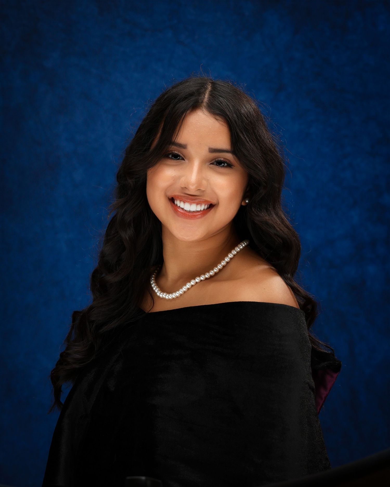 Portrait of someone in a black off-shoulder dress and pearl necklace smiling against a dark blue background.