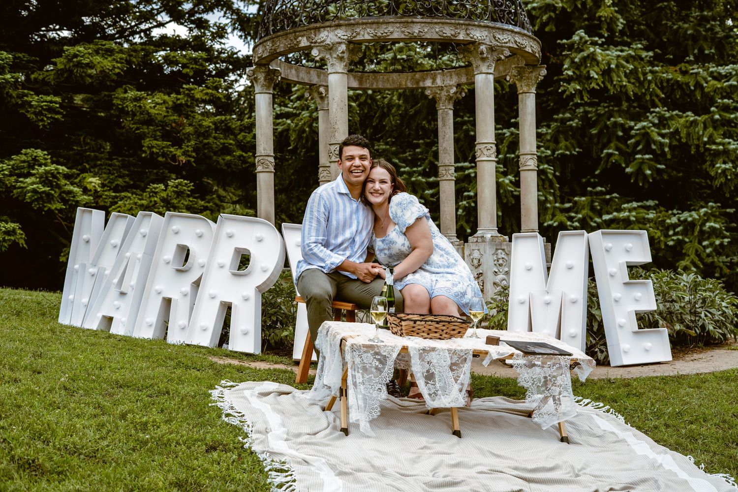 A romantic marriage proposal setup with large white MARRY ME letters and an elegant gazebo in a lush garden setting.