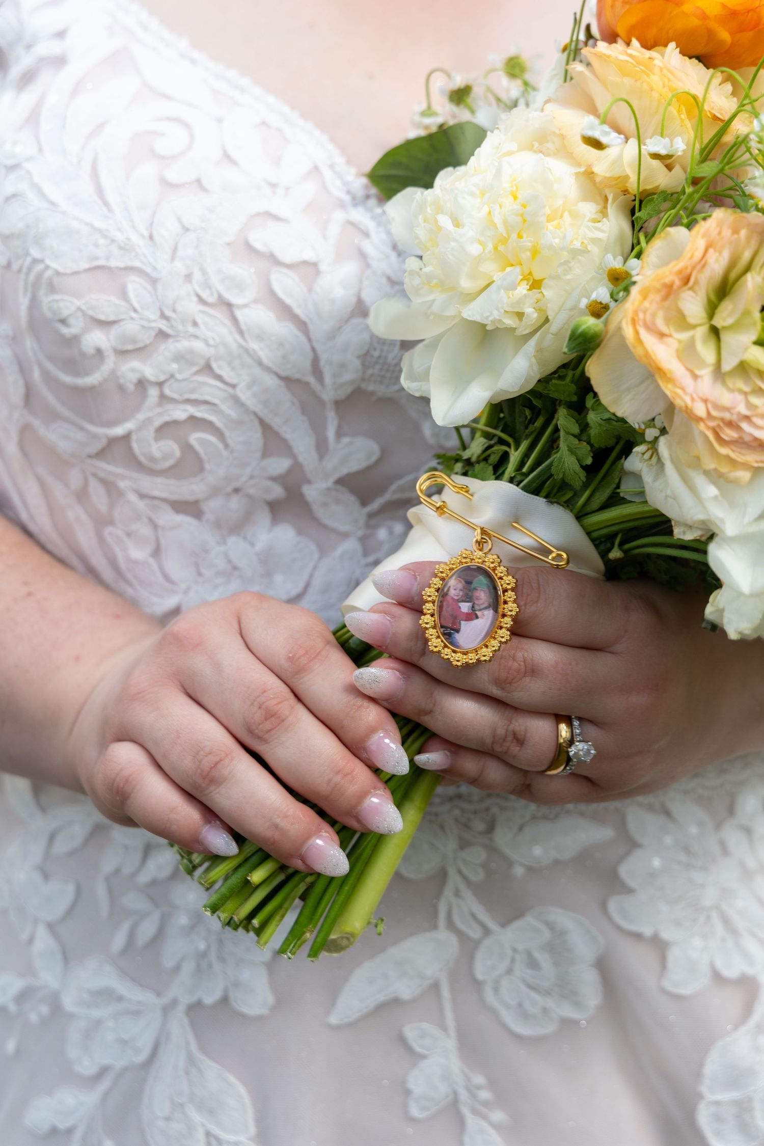Bride in a lace gown holds a peach and white rose bouquet with a gold oval photo charm pinned to the ribbon-wrapped stems.