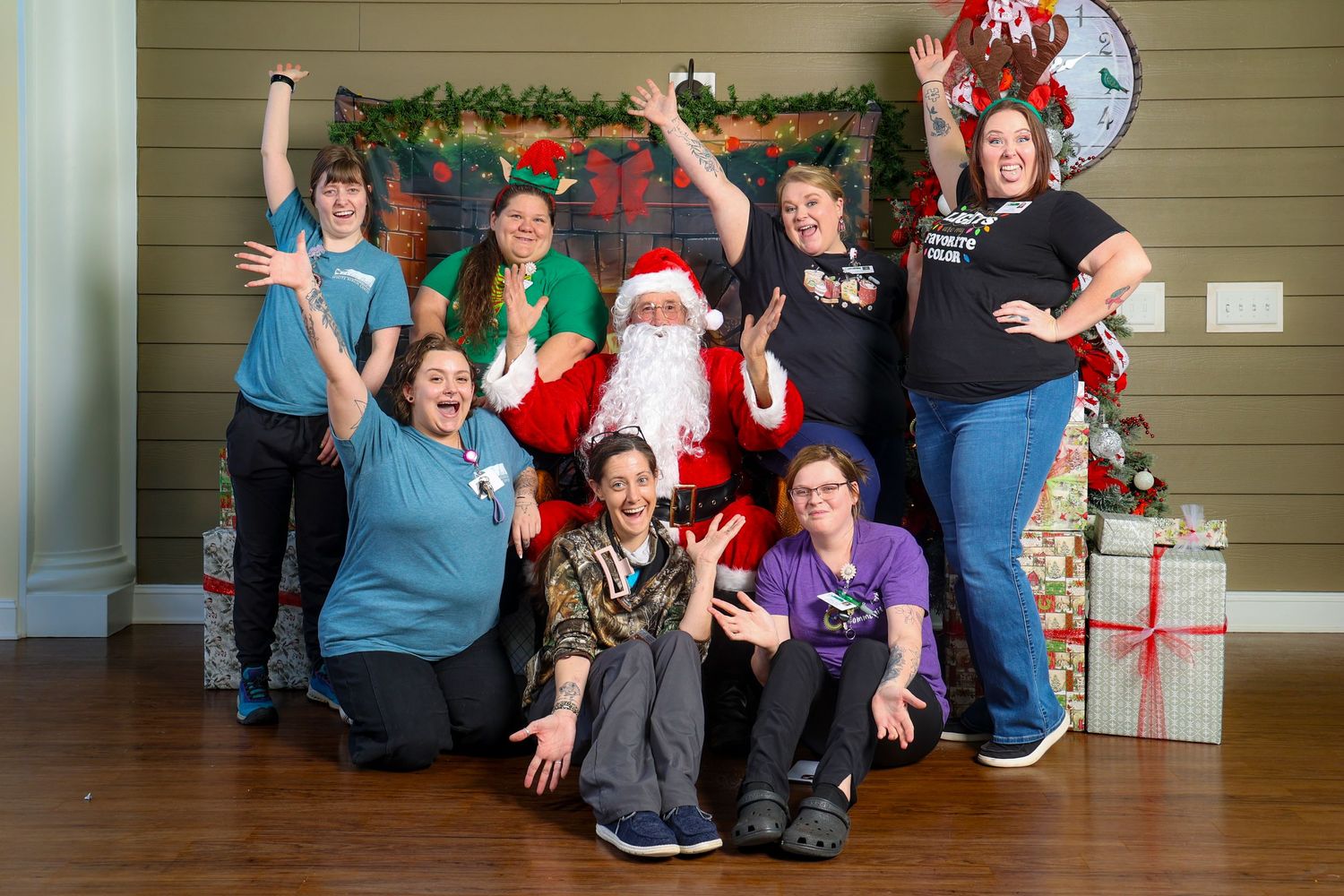 A festive holiday group photo with Santa Claus surrounded by people posing cheerfully in front of a decorated wall.