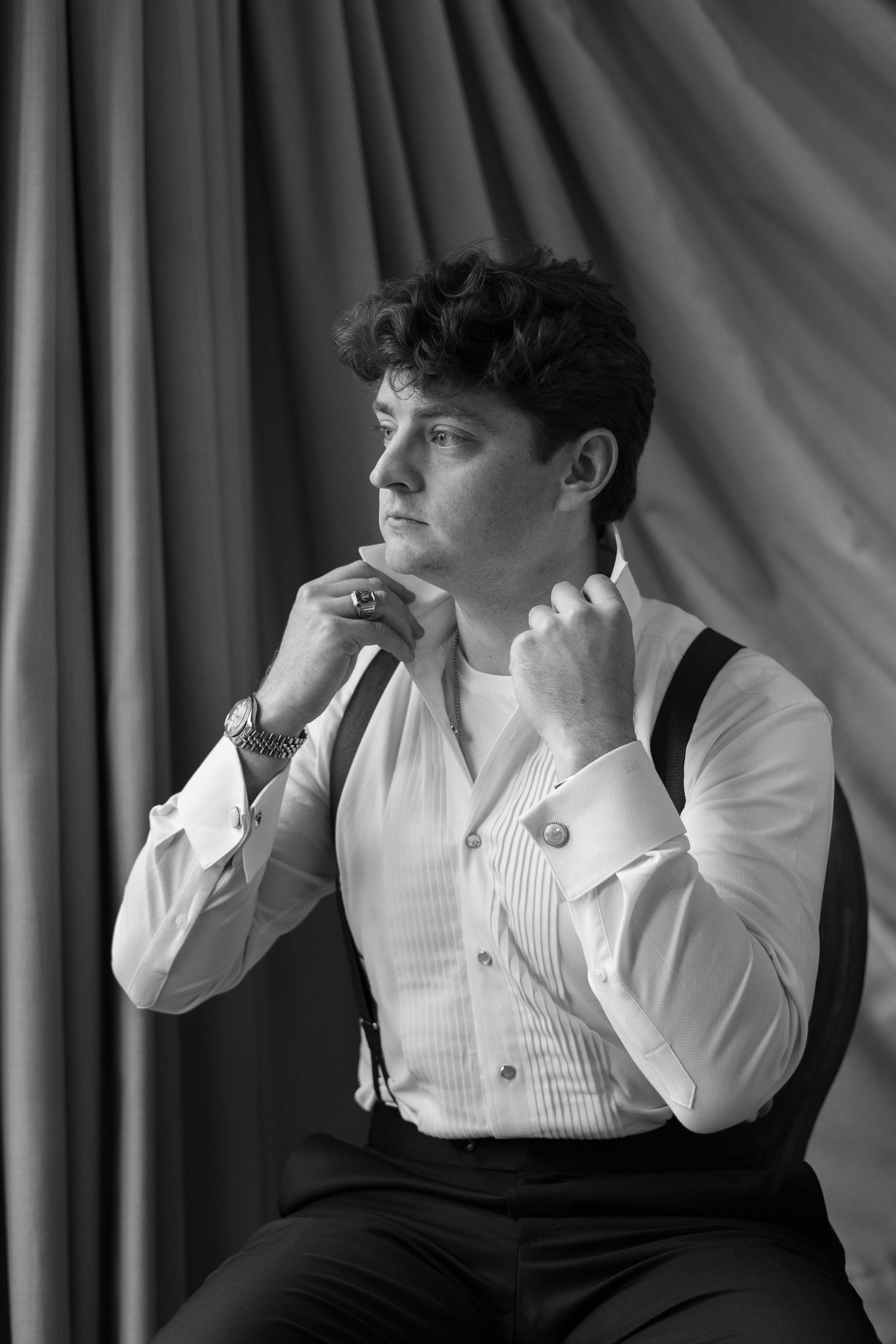 Black and white portrait of a man with curly hair in a pleated dress shirt and suspenders, seated and gazing aside against a draped curtain.