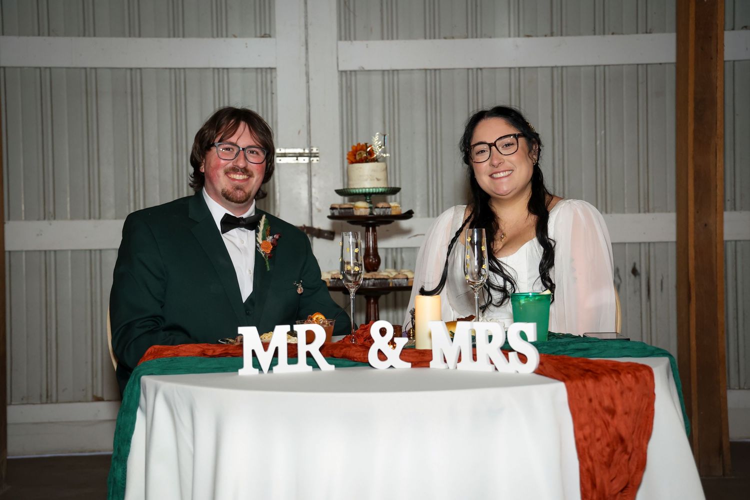 A couple sits at their wedding reception table with a small tiered cake and 'MR & MRS' sign displayed in front.