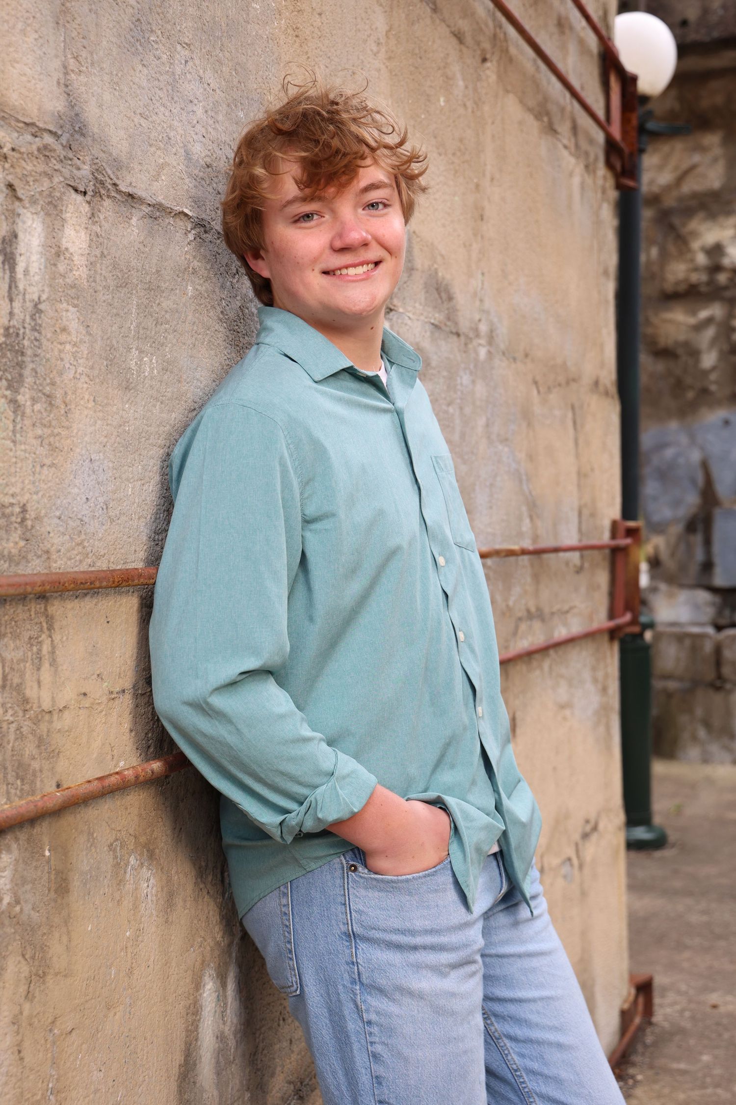 Person wearing a light blue button-down shirt and jeans leans against a textured concrete wall in a casual pose.