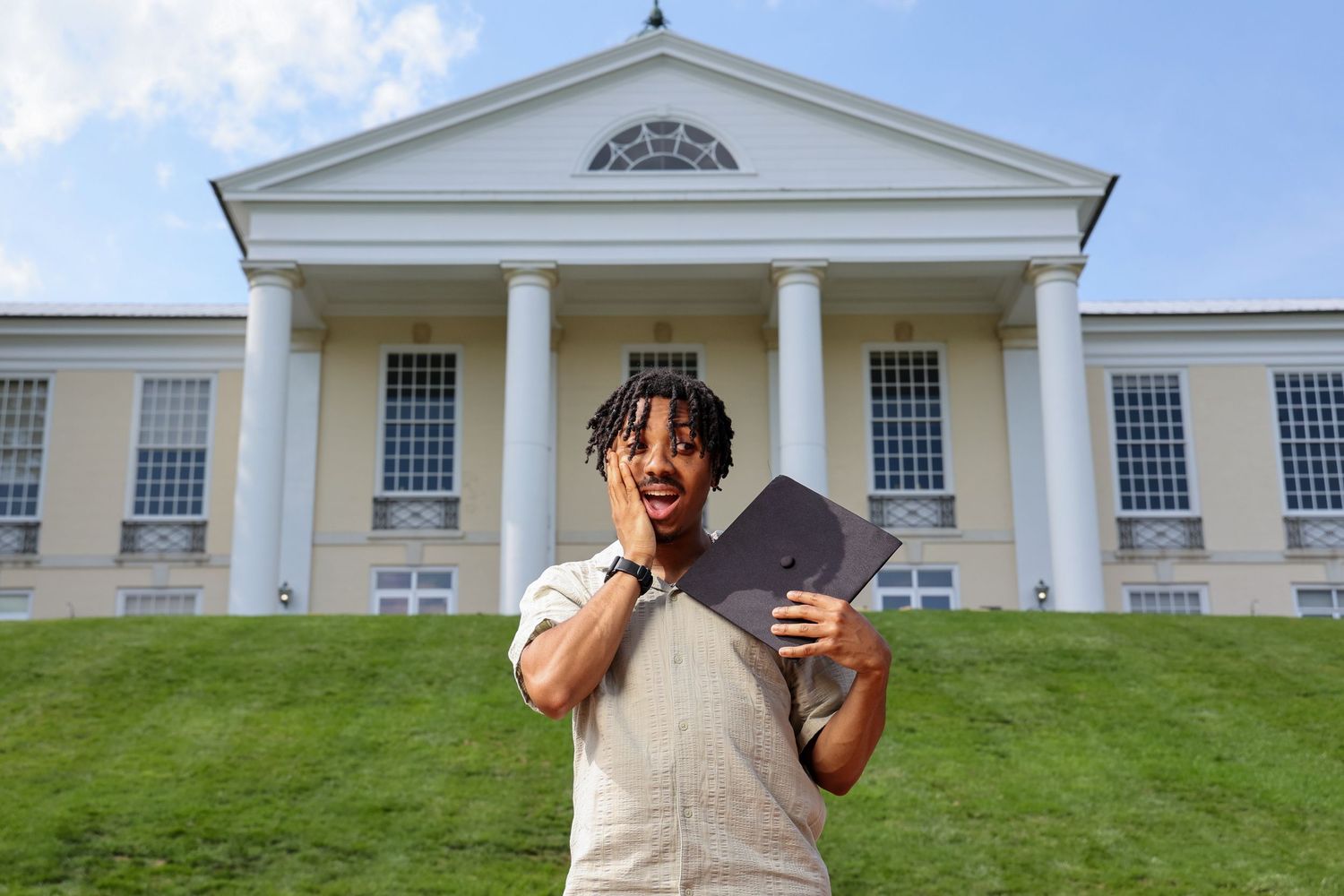 Student holds a book while standing in front of a stately white building with columns and a triangular pediment.