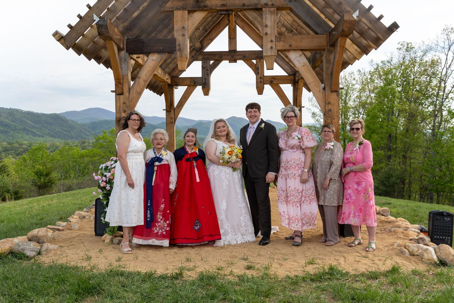 Wedding party of bride, groom and guests, some in traditional Korean hanbok, pose under a rustic wooden pavilion with mountain views.