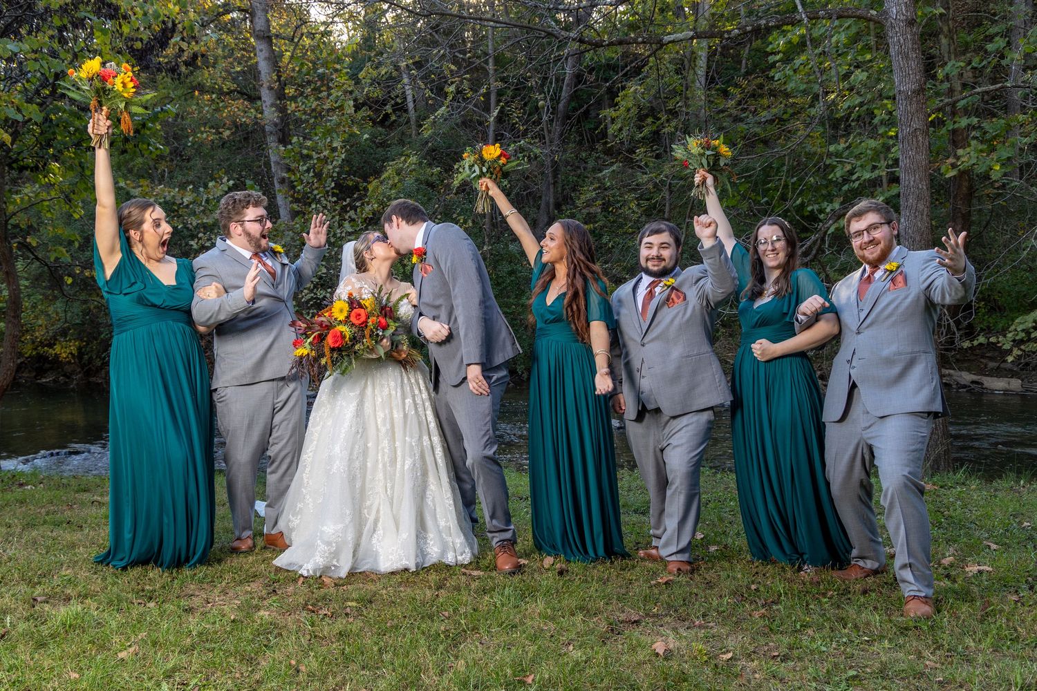 Wedding party in gray suits and teal dresses poses together joyfully outdoors in a natural setting.