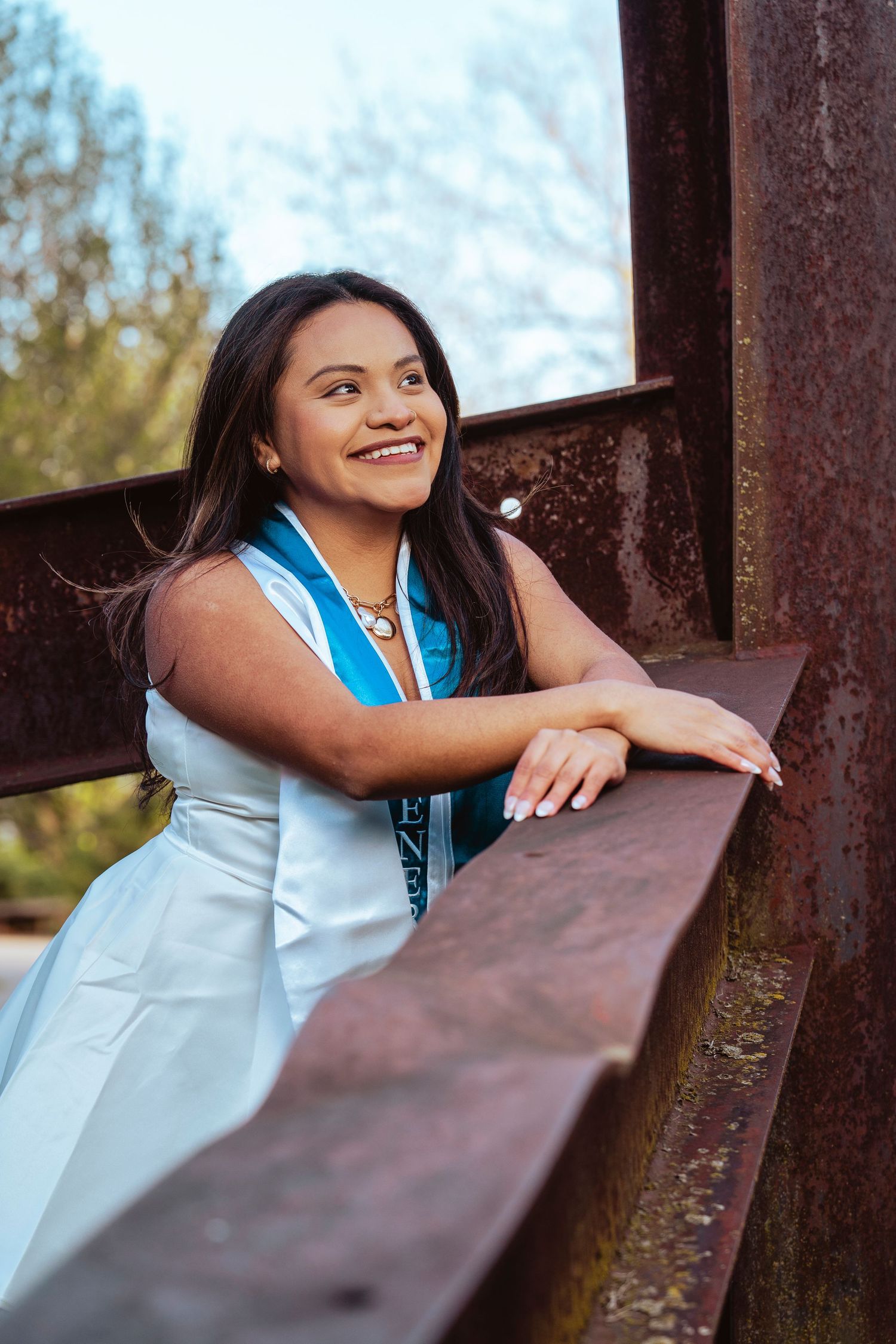 A graduate in white and blue regalia leans on a rustic wooden railing while smiling during an outdoor photo session.