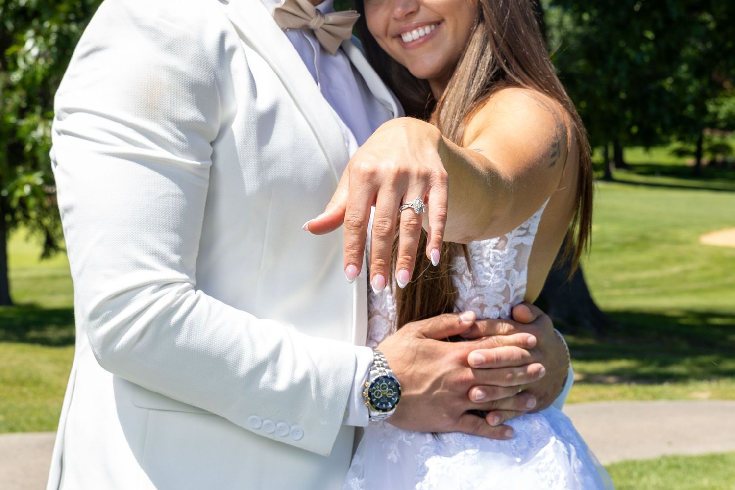 A couple in wedding attire displays an engagement ring outdoors in a sunny park setting.