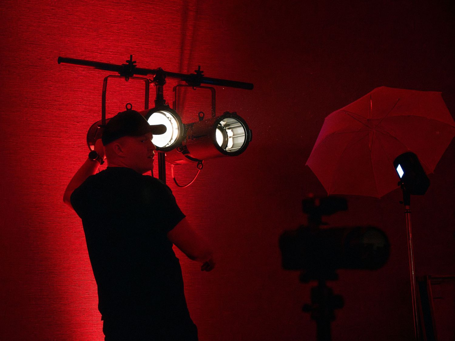 Silhouette of a camera operator filming against dramatic red lighting with studio equipment and umbrellas visible.