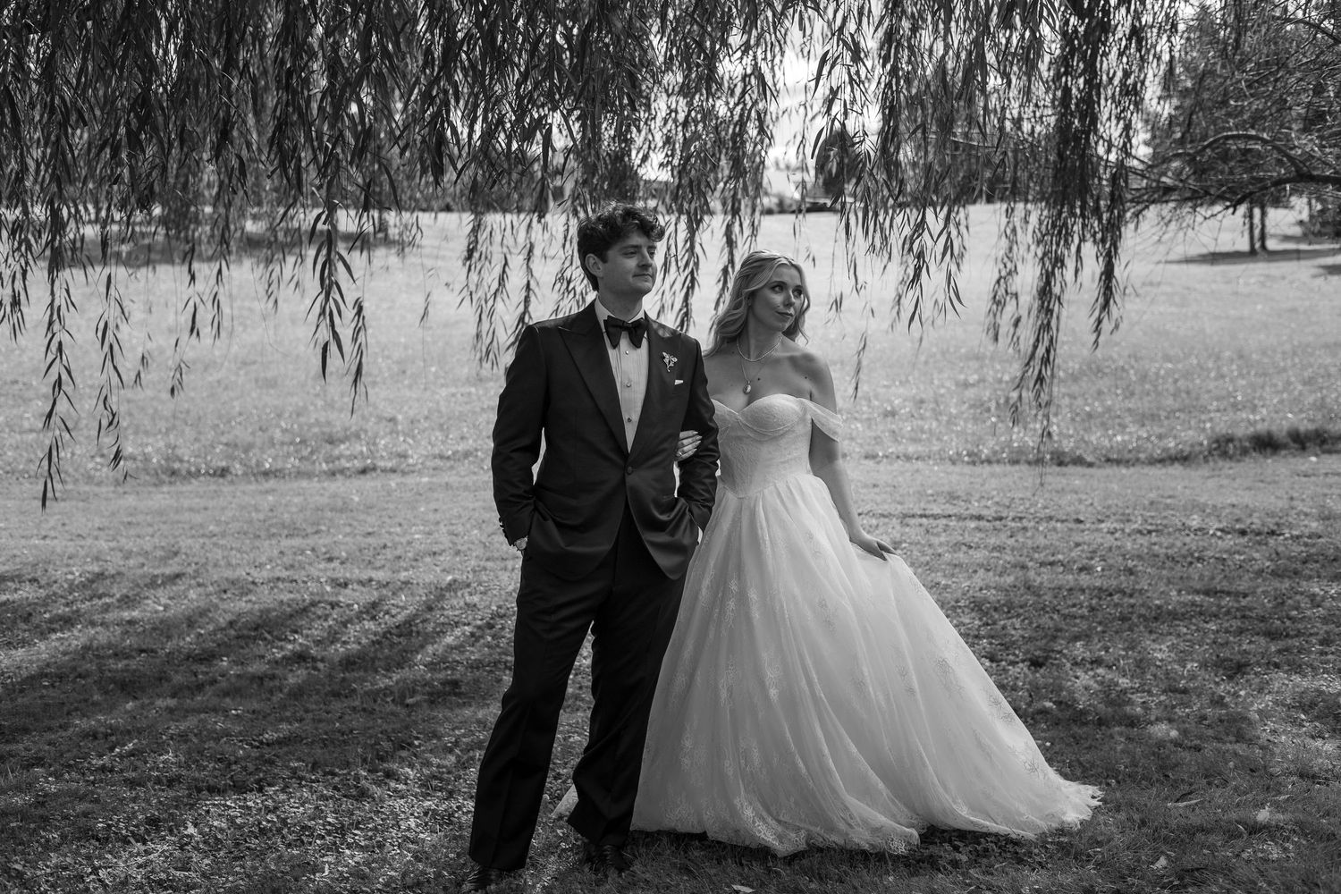 Black and white photo of a bride in a strapless ballgown and a groom in a tuxedo standing under willow branches in a field.