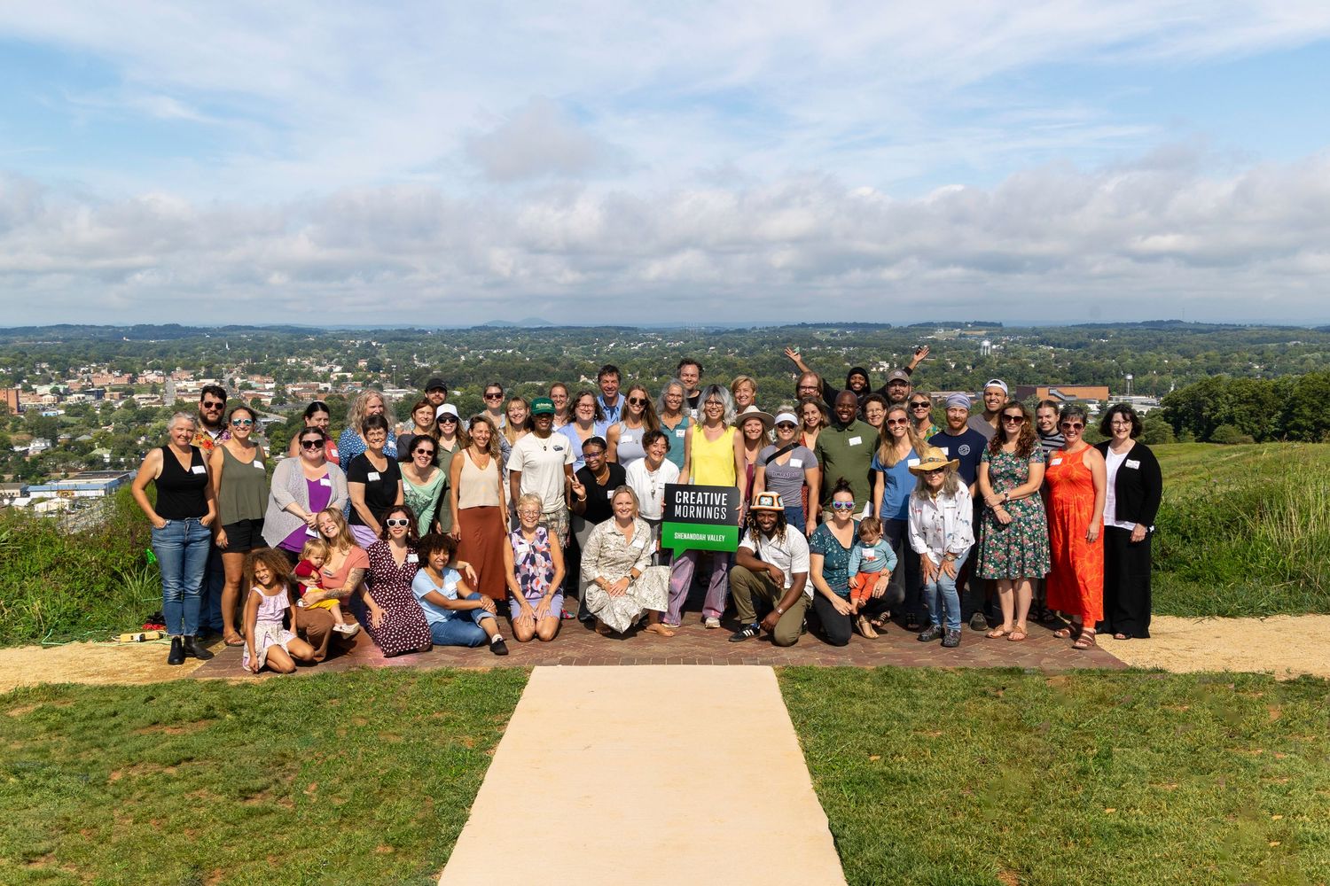 Large panoramic group photo of people gathered outdoors on a grassy hilltop with scenic mountain views in the background.
