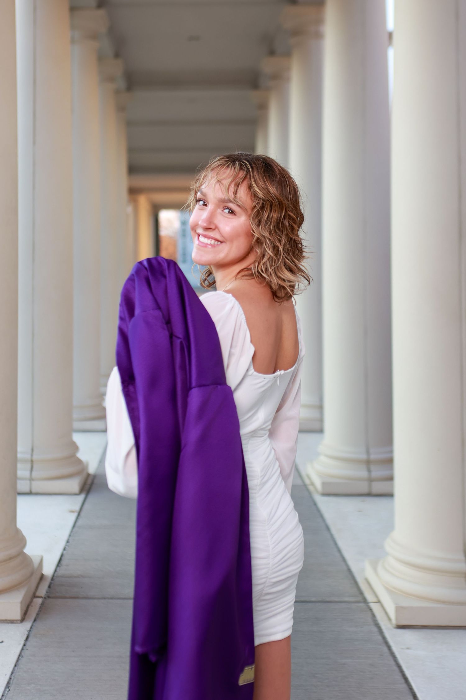 A person in a white dress poses playfully with a purple graduation gown in a columned hallway.