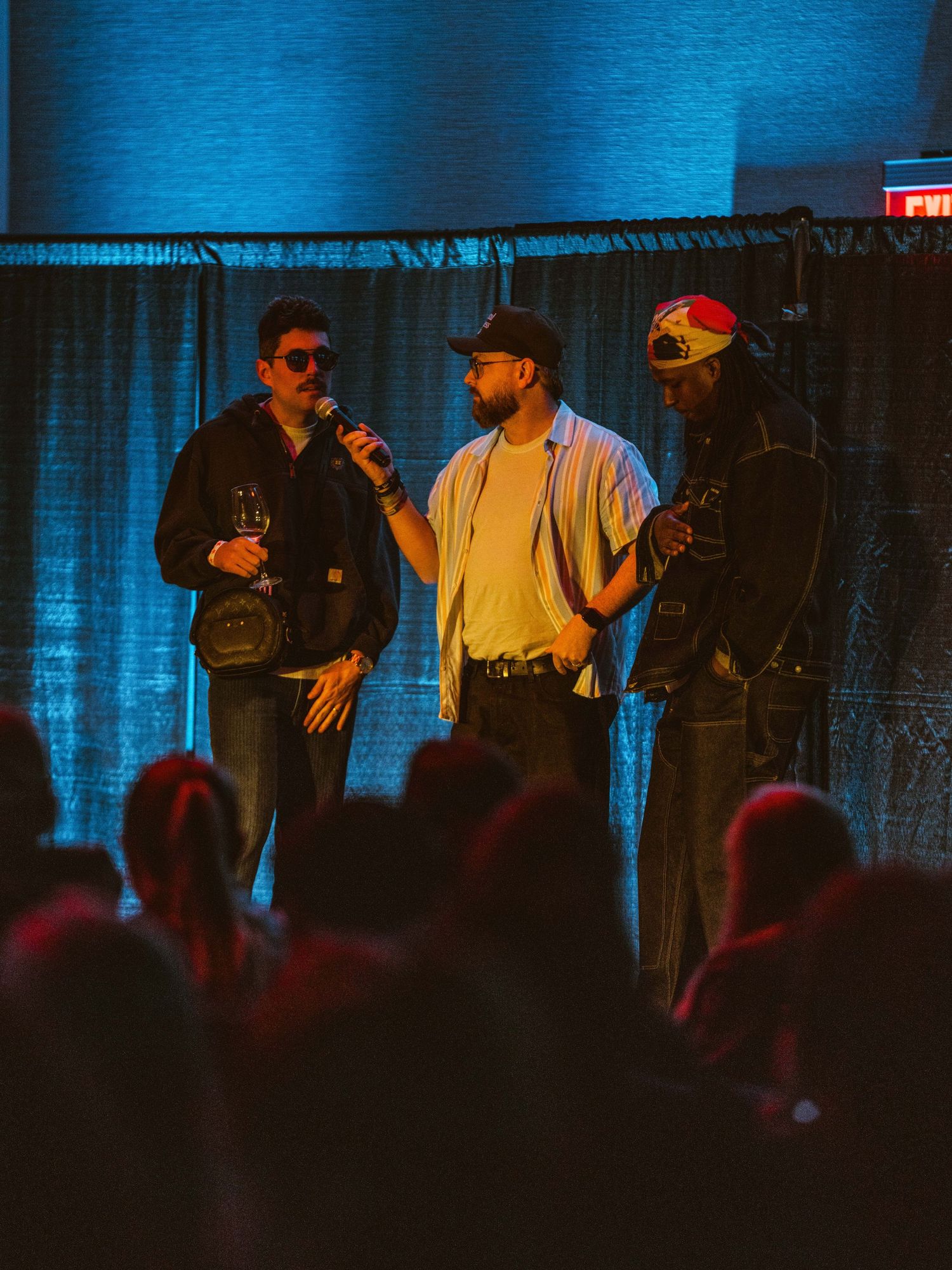 Three performers on stage with blue lighting and silhouetted audience members visible in the foreground during a live show.