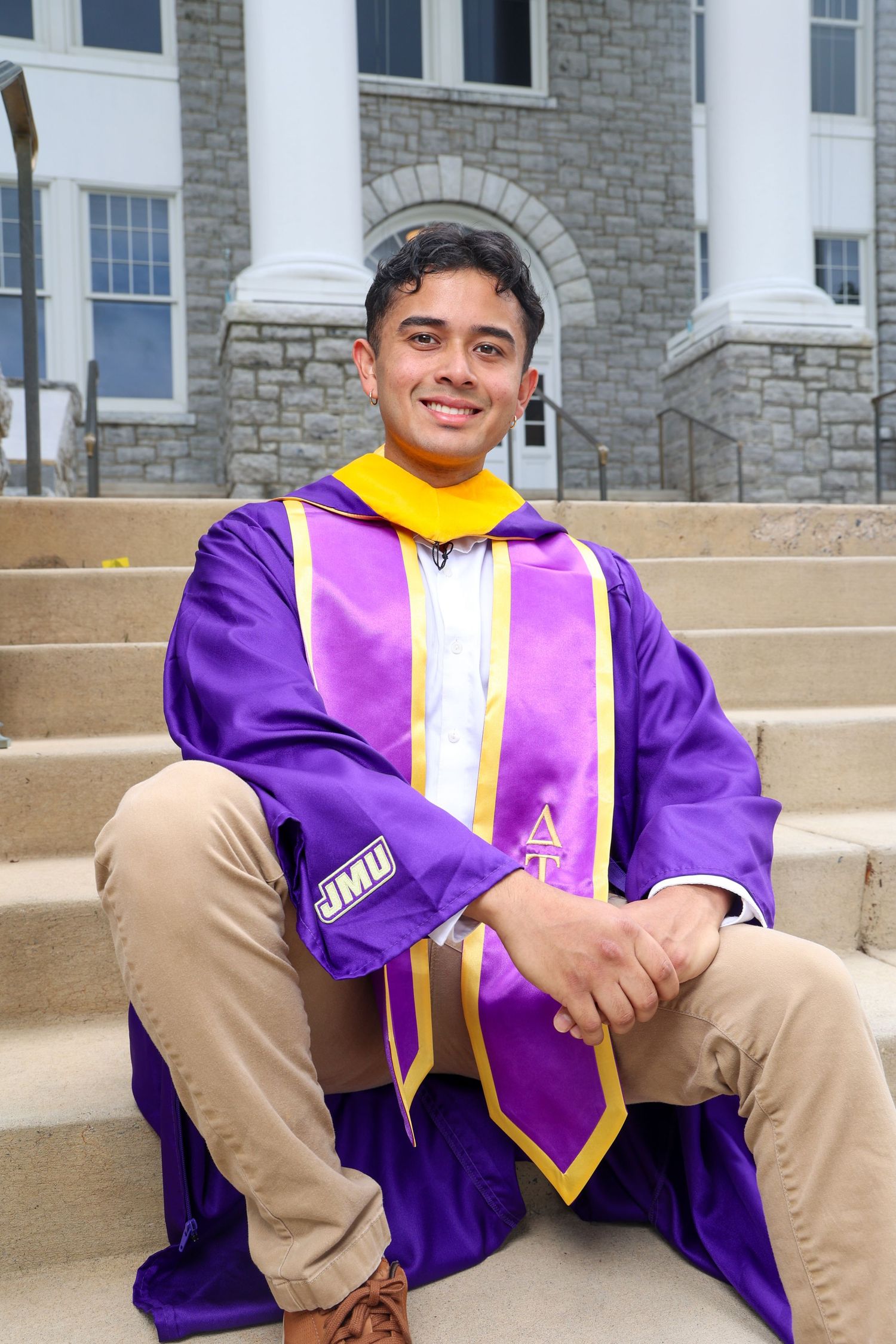 Graduate seated on stone steps wearing purple and yellow academic regalia.