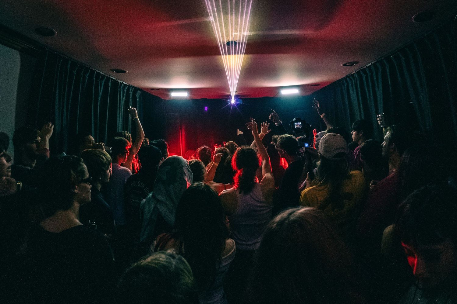 A crowded nightclub dance floor with silhouetted people under red lighting and a bright beam from above.