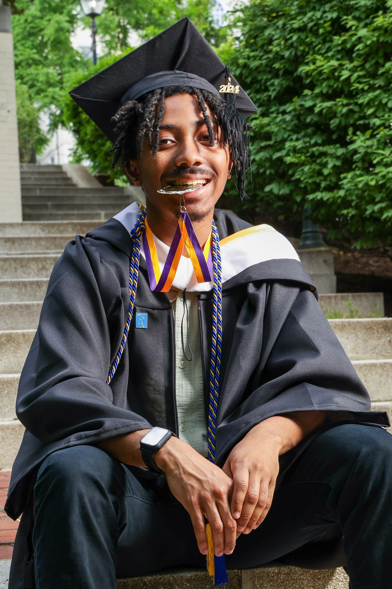 Graduate in cap and gown wearing honor cords sits smiling on stone steps during commencement ceremony.