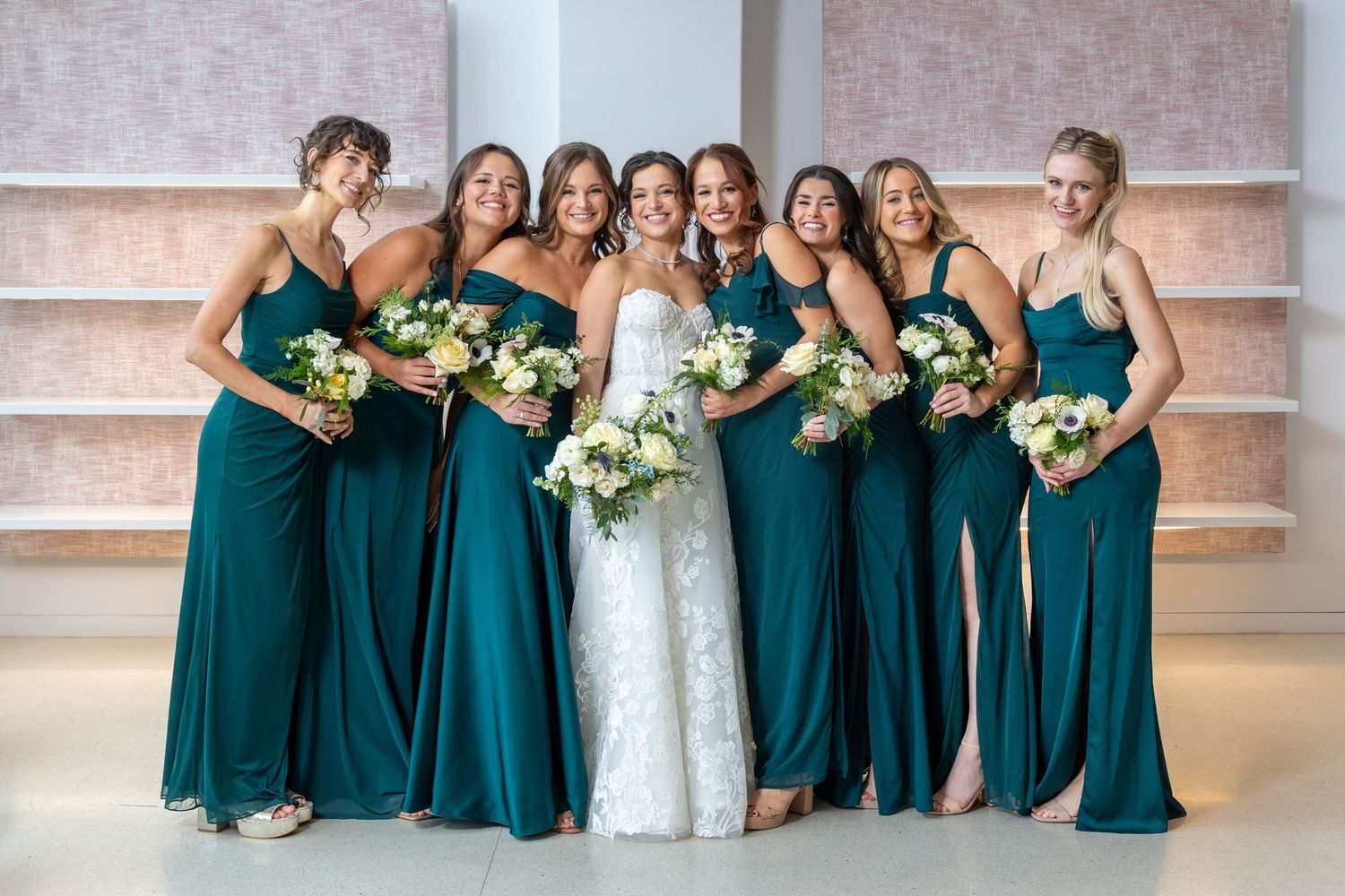 Wedding party in elegant emerald green dresses poses together while holding white floral bouquets.