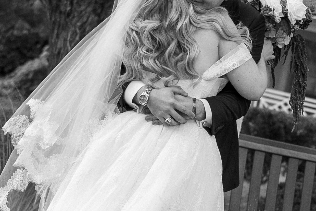 Black and white photo of a bride in a lace veil and gown embracing her groom in a dark suit beside a wooden bench and flowers.