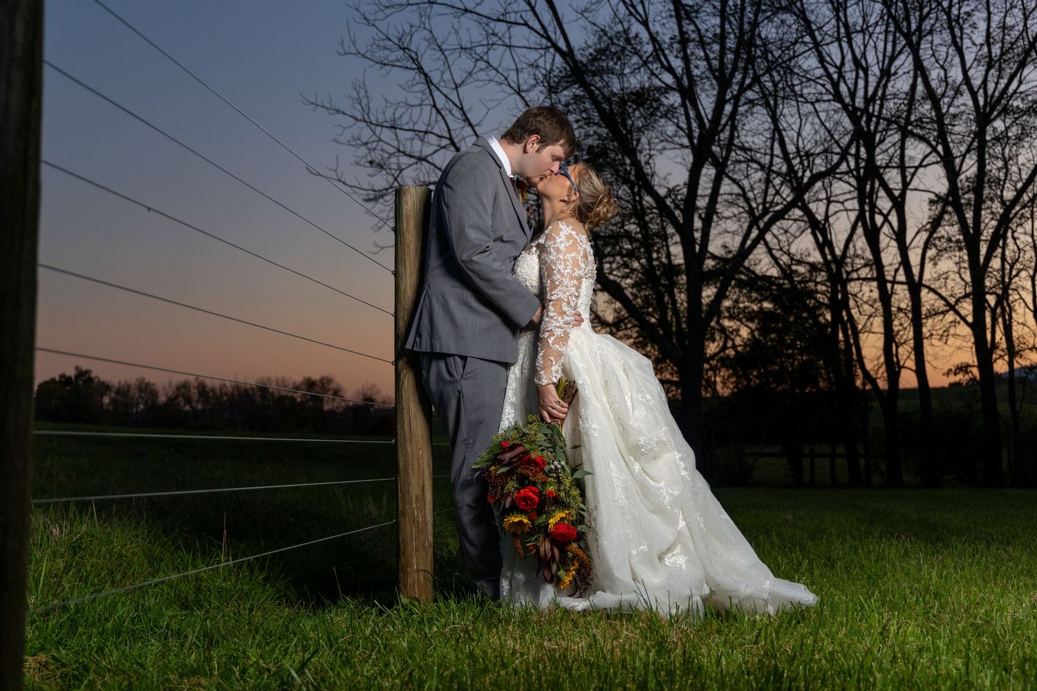 A couple in wedding attire poses romantically at sunset beneath power lines in a grassy field.