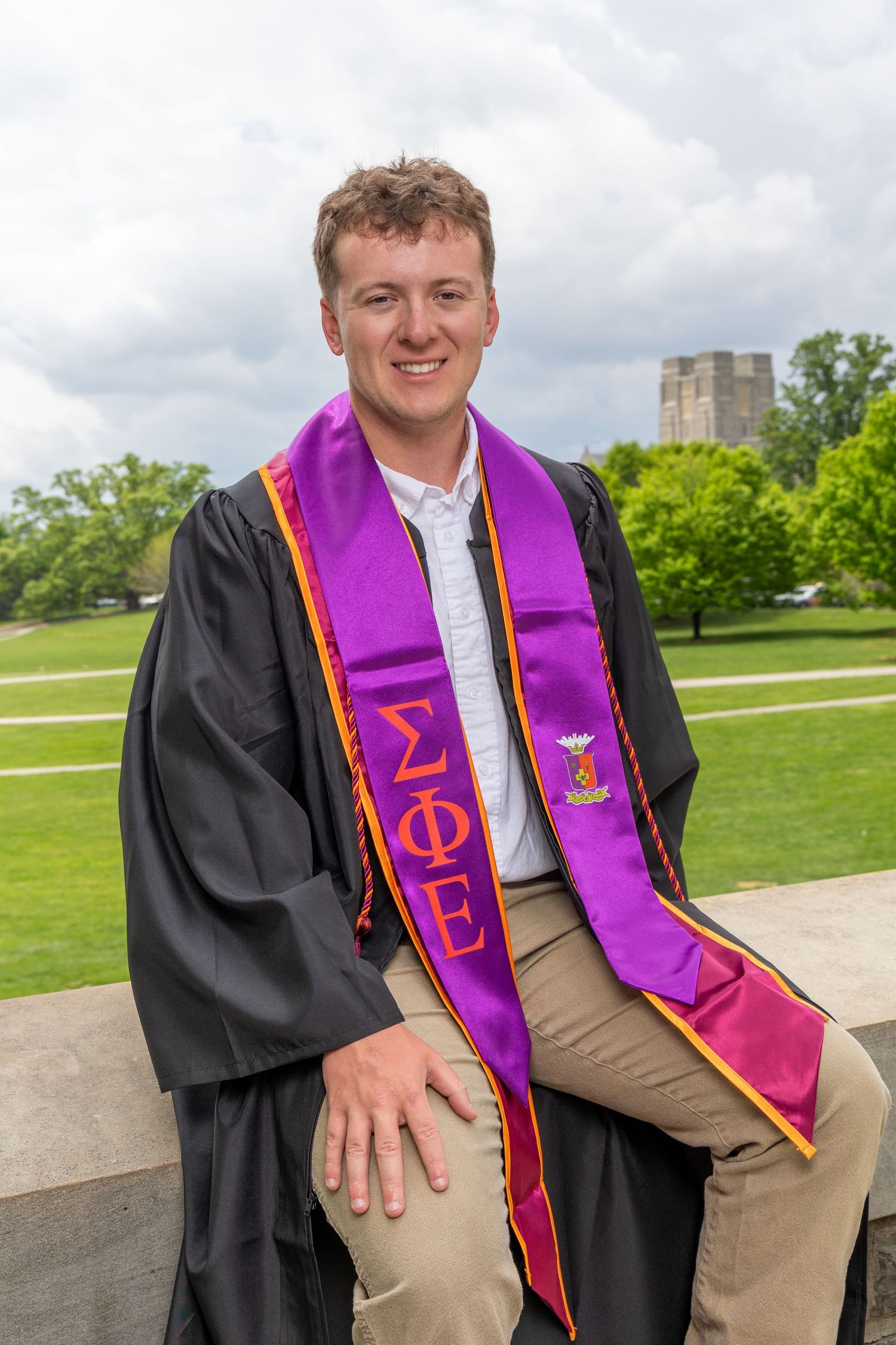 Graduate wearing black gown and purple stole sits on stone steps during commencement photo session outdoors.