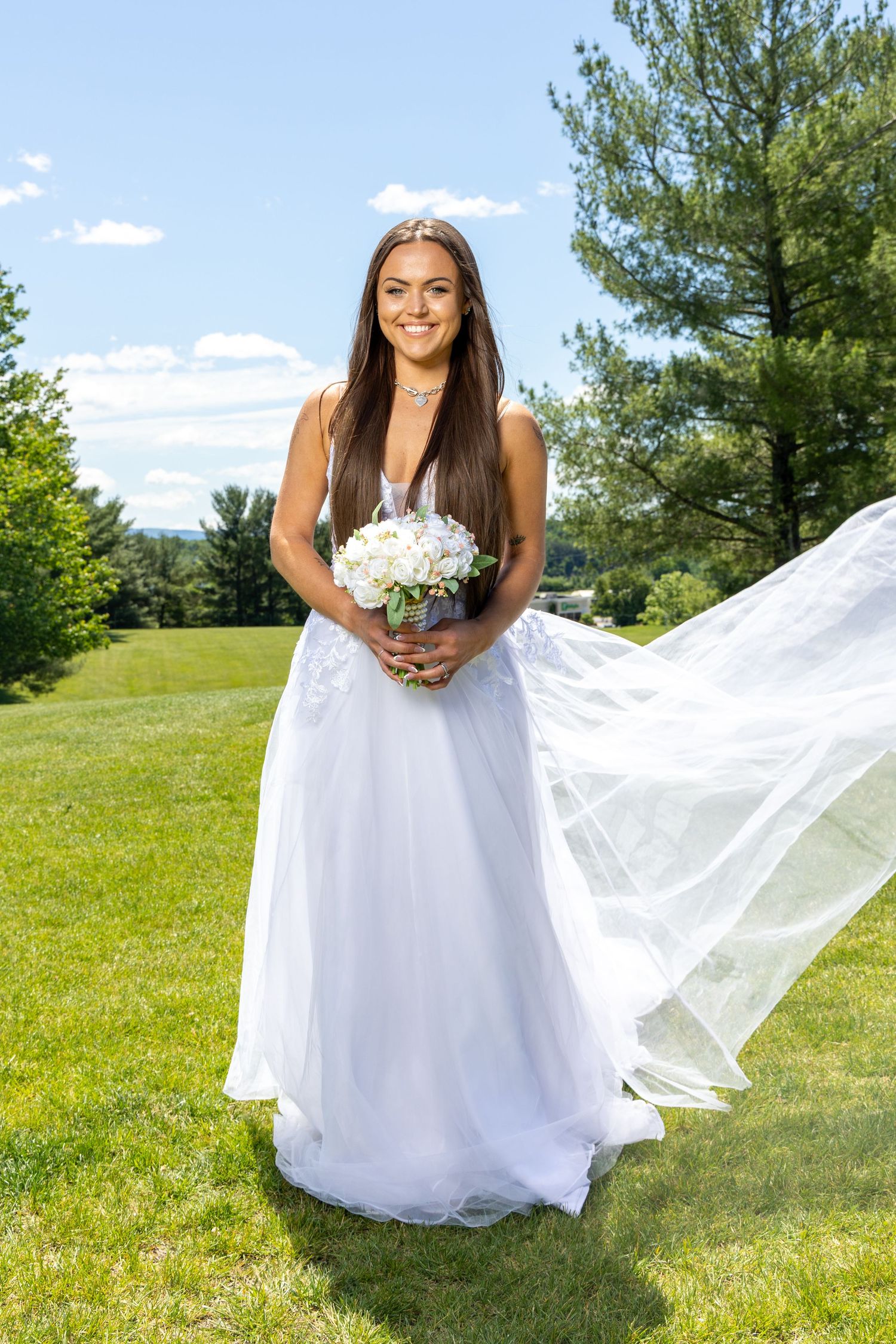 A bride in a flowing white gown holds a bouquet while her veil billows in the wind on a sunny grassy hillside.