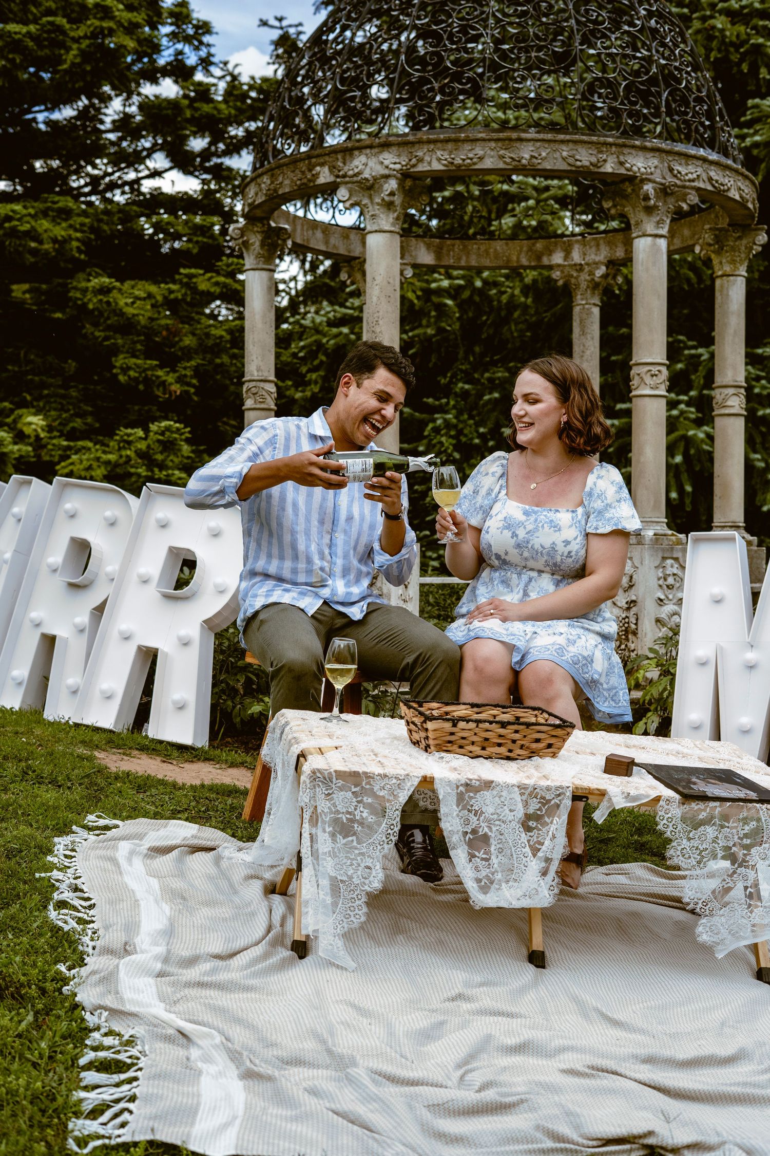 A couple shares a picnic and laughs together on white Adirondack chairs in a garden setting.