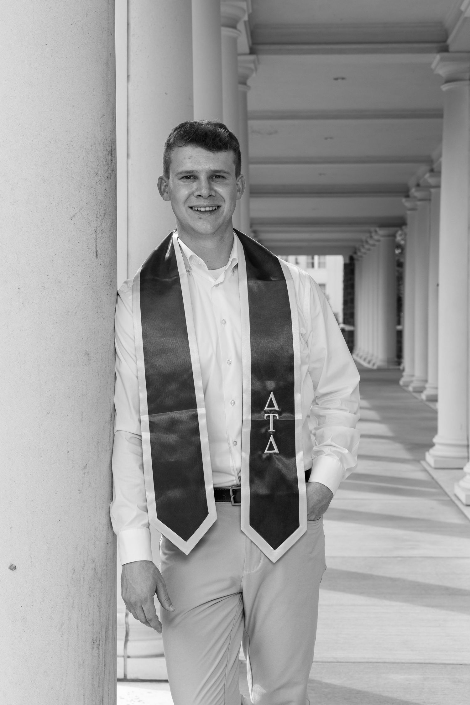 Graduate in formal attire and black stole standing in columned hallway.