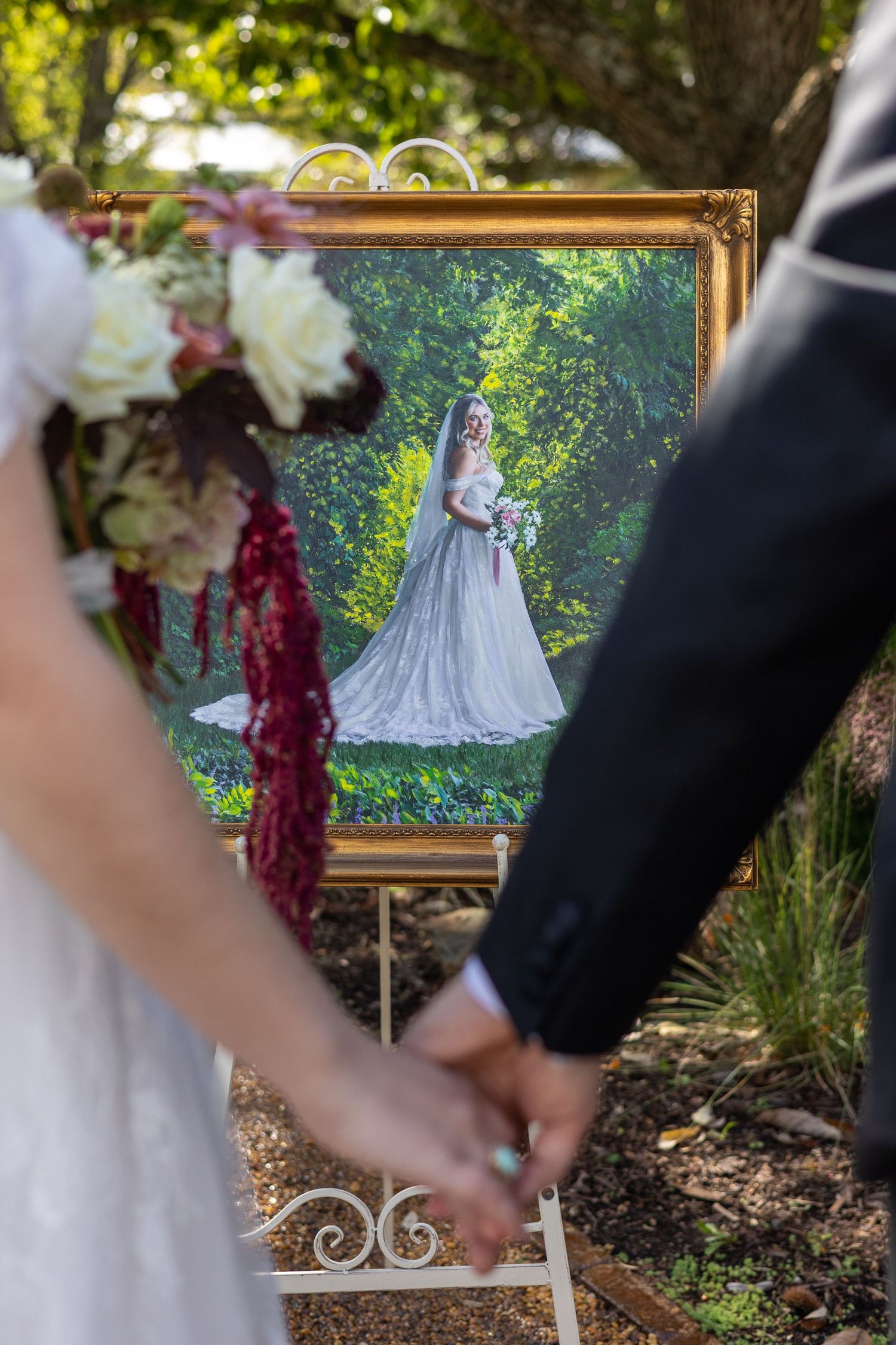 Couple holding hands beside a gold-framed bridal portrait on an easel, with a white and burgundy floral bouquet in a garden wedding setting.