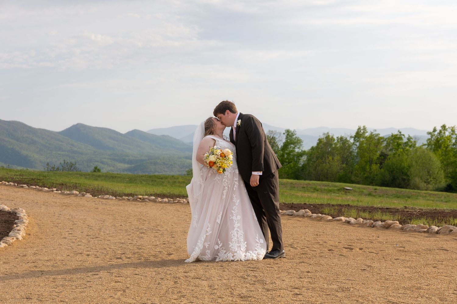 Bride in a white gown holding a yellow bouquet kisses her groom in a dark suit on a mountaintop wedding ceremony site with scenic views.