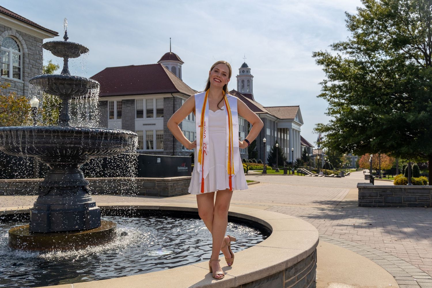 A person walks along the edge of a decorative fountain at a public plaza on a sunny summer day.