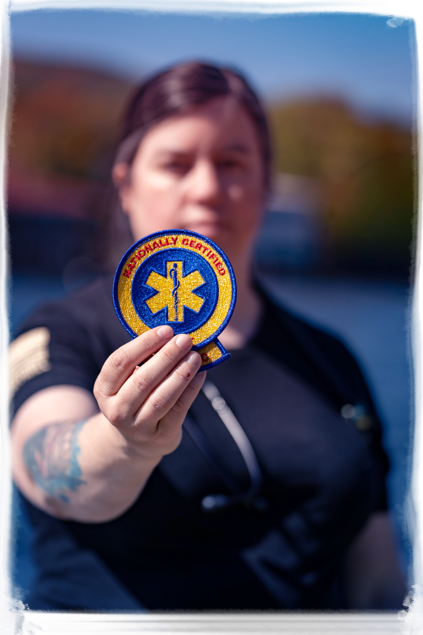 Emergency medical technician holding up a blue and gold EMT patch against blurred outdoor background.