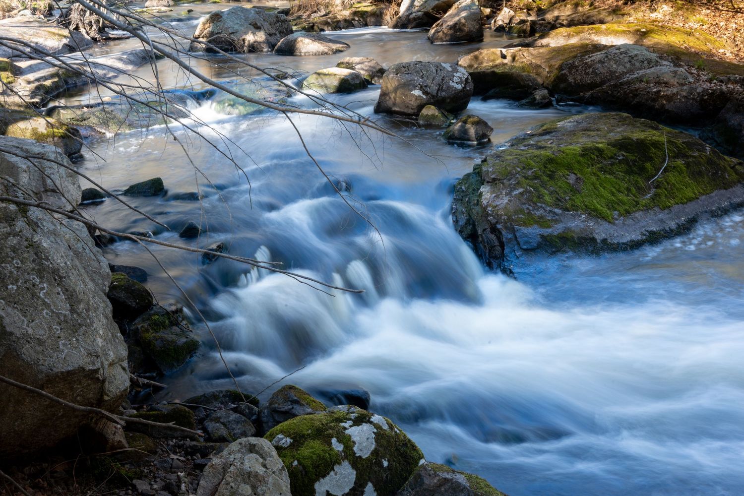 A rushing mountain stream flows over mossy rocks with silky smooth water effect captured by long exposure photography.