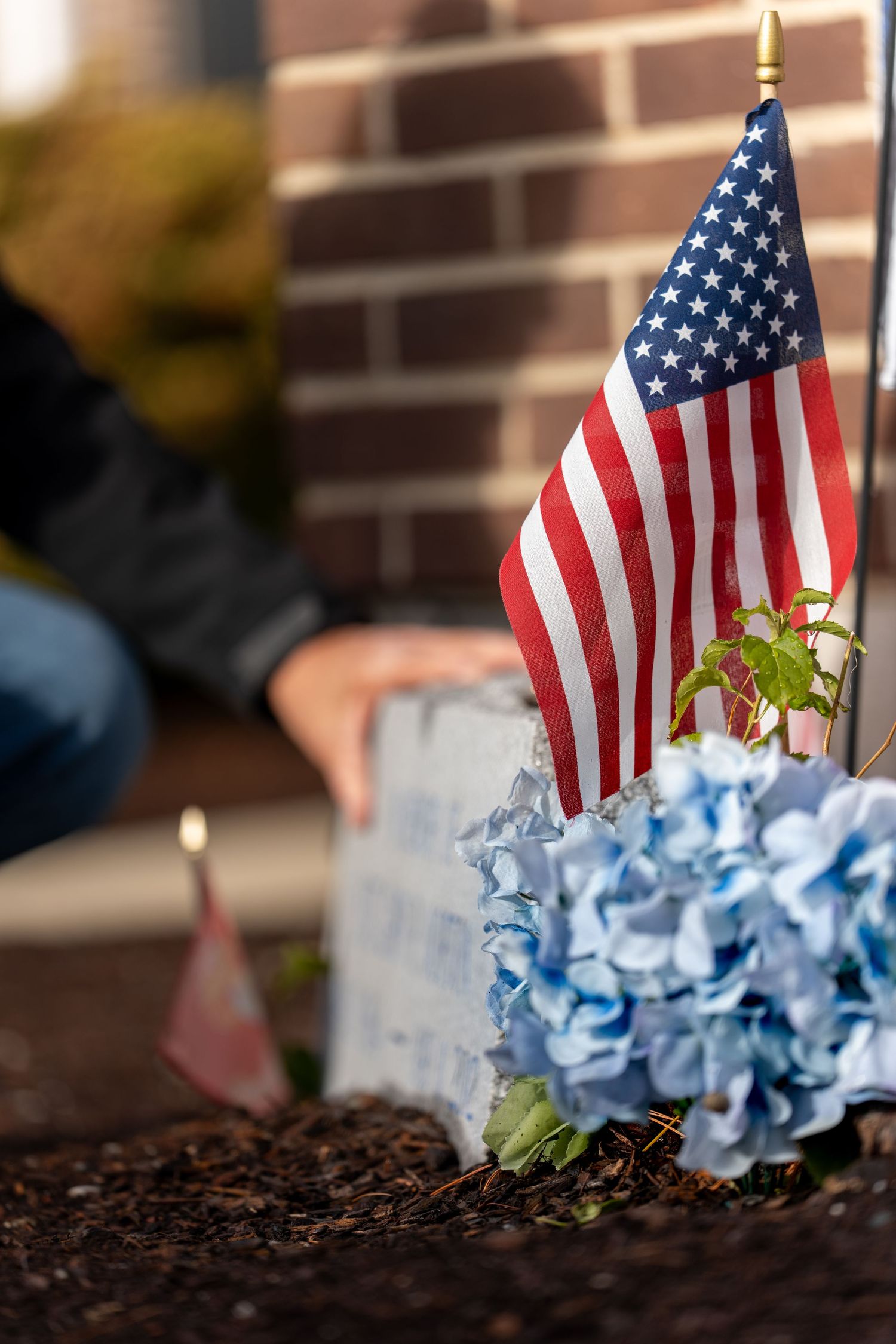 Small American flag planted among blue hydrangeas and soil in a memorial or garden setting.