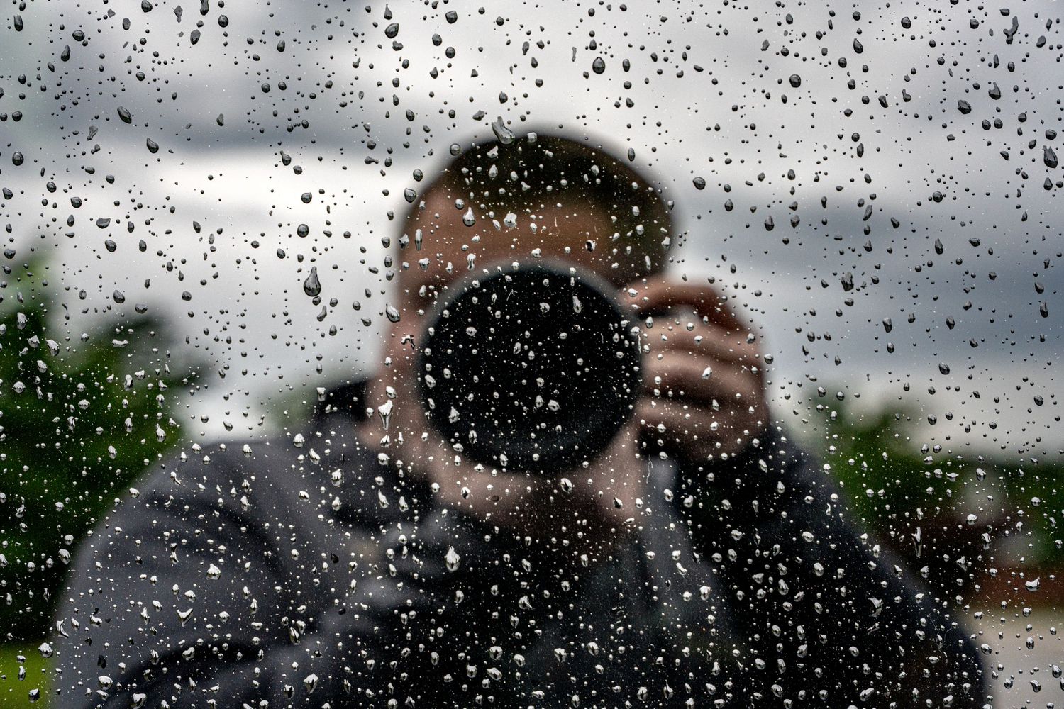 Photographer capturing self-portrait through rain-covered glass window on overcast day.